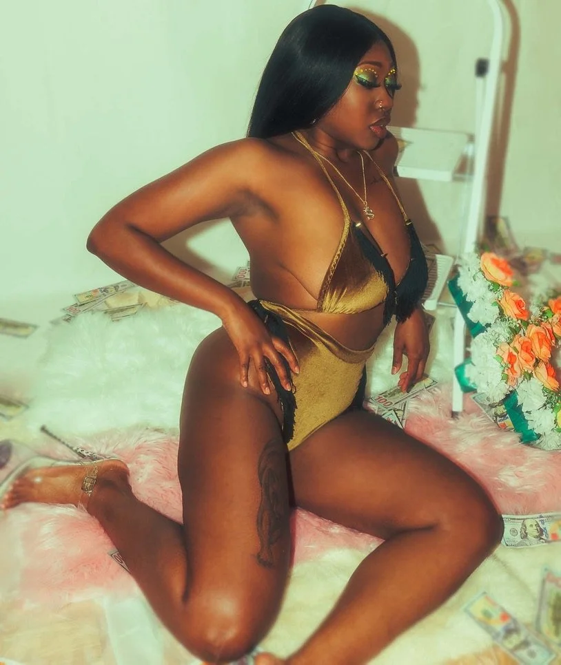 A woman with long black hair and shiny makeup, wearing a gold and black lingerie outfit, kneeling on a bed surrounded by scattered money and a bouquet of flowers.