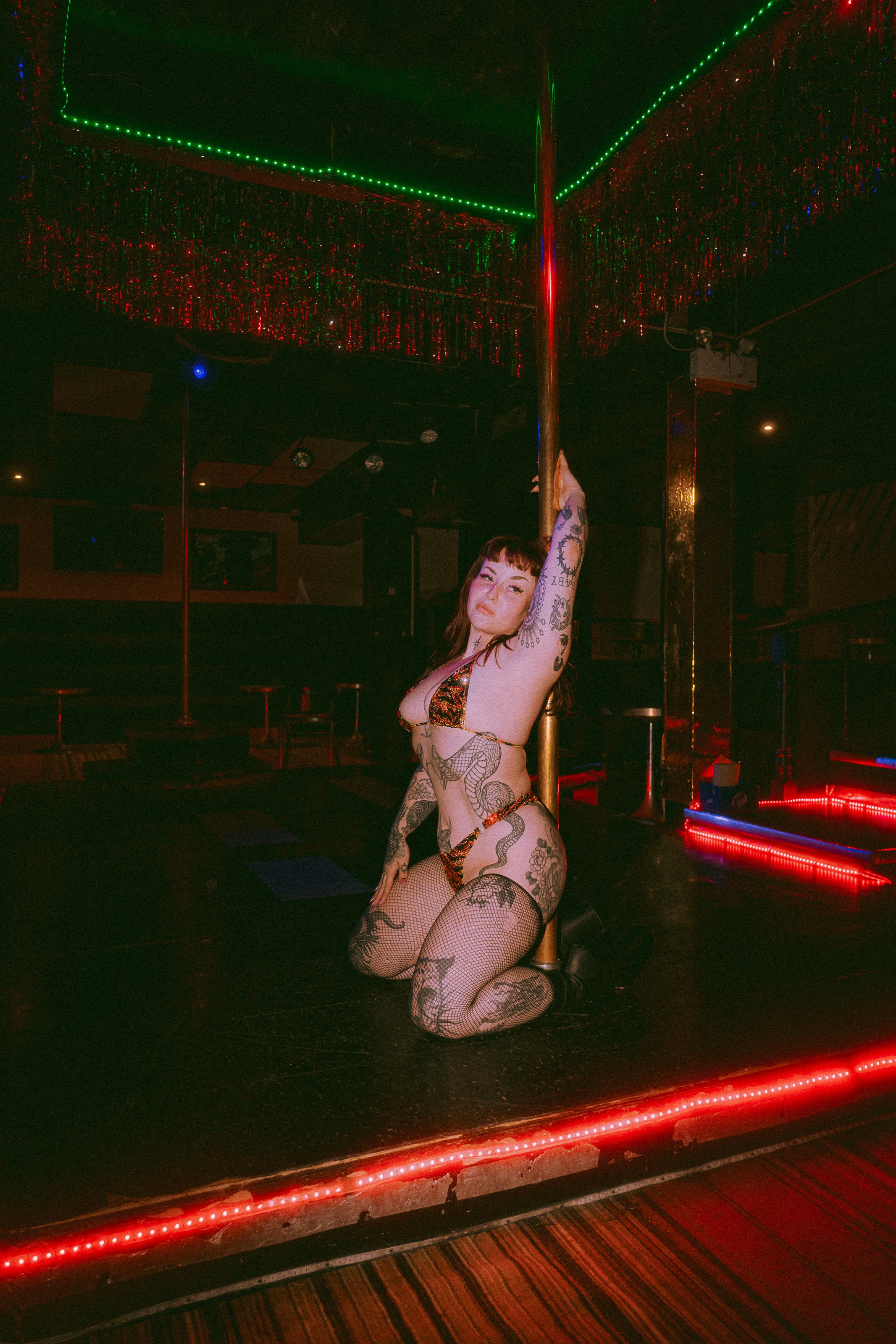 A woman with tattoos on her arms and legs is kneeling on a stage with a pole, wearing a leopard print lingerie set. The stage has red and green neon lights, and a mirrored ceiling with red and green LED lights.