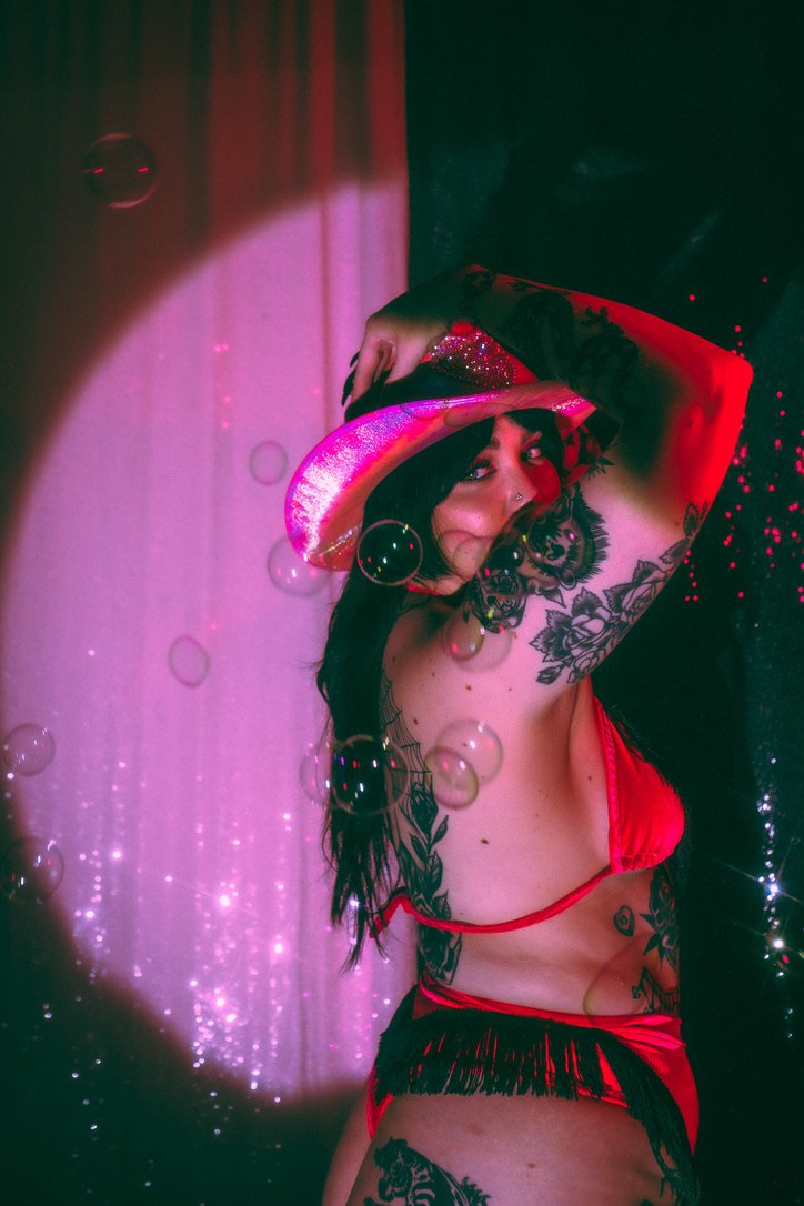 A woman with tattoos wearing a red cowboy hat, red lingerie, and black fringe shorts, posing with her arm behind her head against a backdrop with pink and black lighting and floating bubbles.