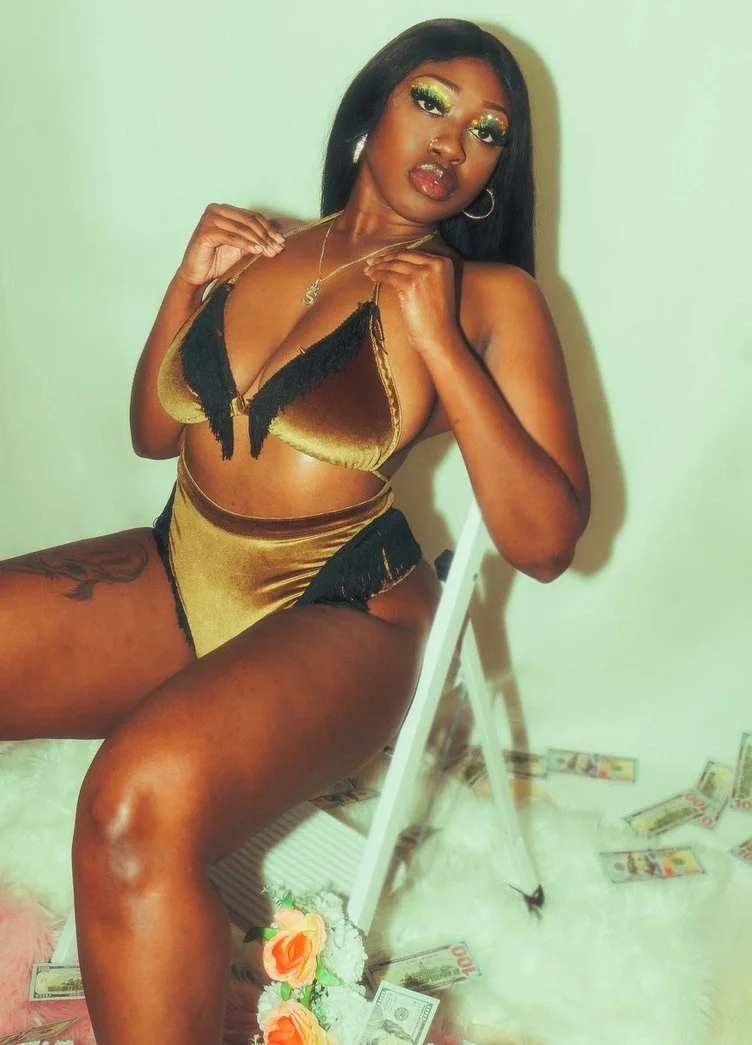 A woman sitting on a white chair, wearing gold and black lingerie, with colorful makeup, jewelry, and surrounded by scattered fake dollar bills on the floor.