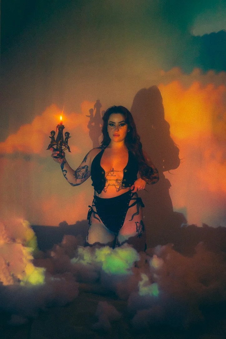A woman with dark hair and tattoos standing in a cloud of smoke or fog, holding a small decorative candle holder with a lit candle, with colorful lighting creating dramatic shadows on the wall behind her.