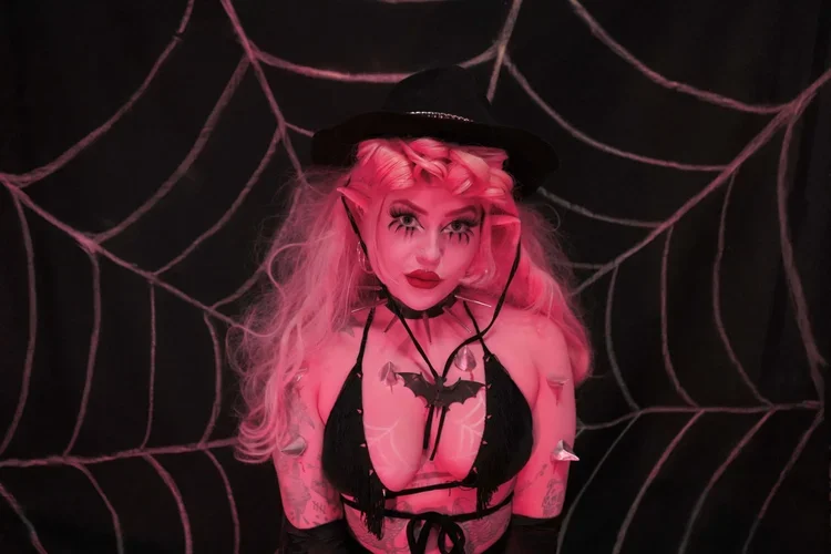 Person dressed in a spider-themed costume with pink hair, makeup, and black hat, posing in front of a black background with large pink web designs.