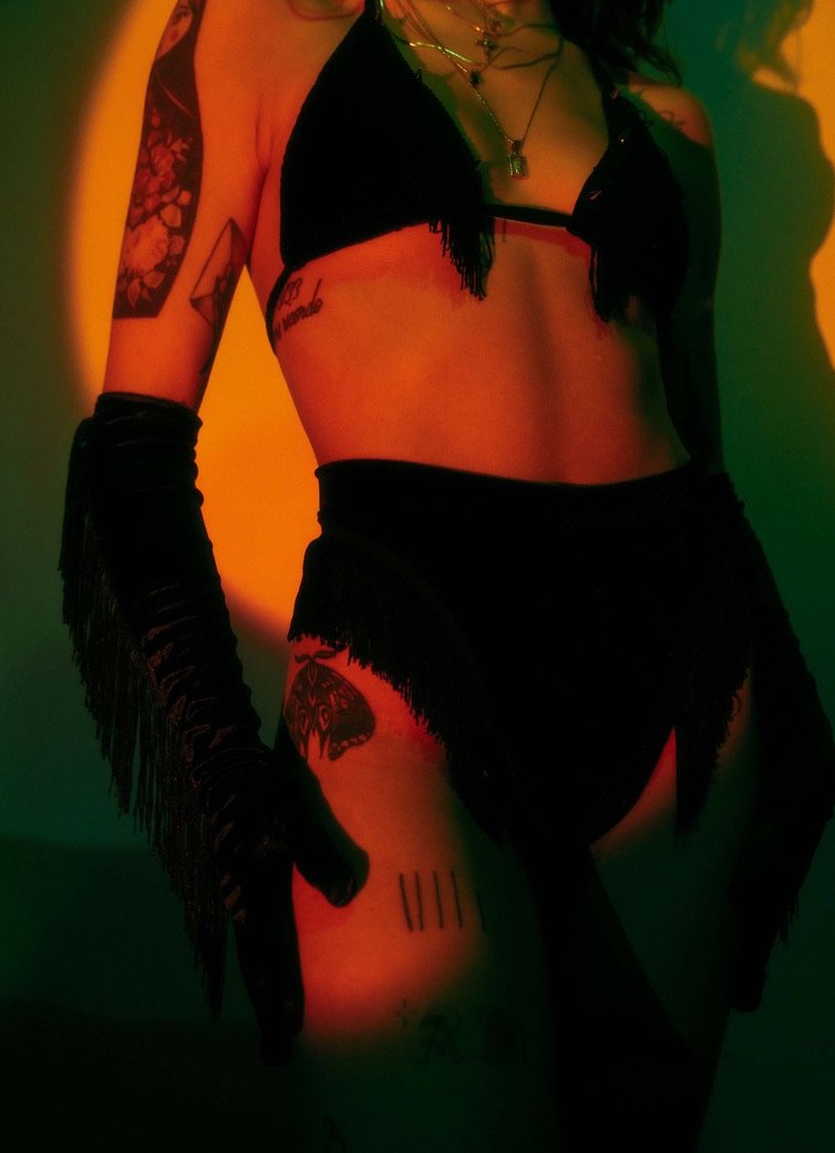 A person wearing black fringe clothing and long gloves, showing her midsection and tattoos, with multicolored lighting.