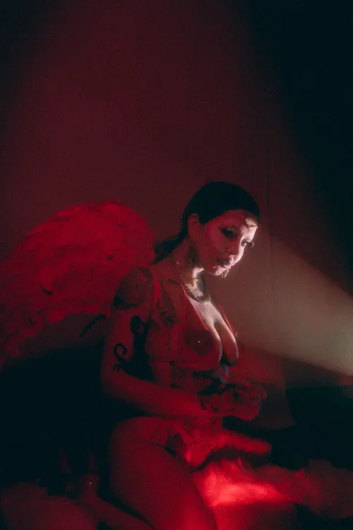 A woman dressed in red with a feathered headpiece and tattoos, sitting in a dark room with red lighting.