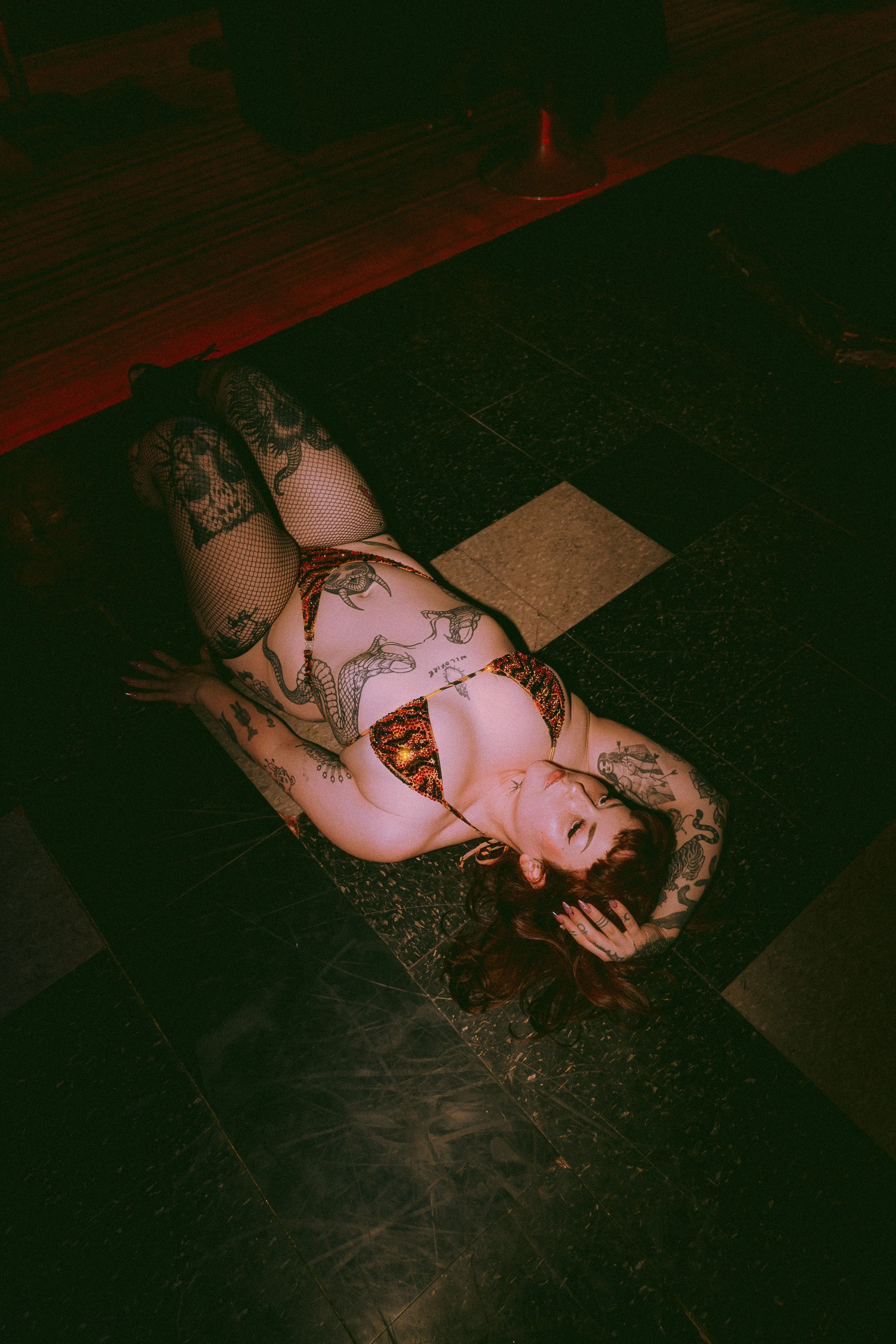 A woman with tattoos, red hair, fishnet stockings, and a leopard print bikini lying on a black tile floor in a dark room.