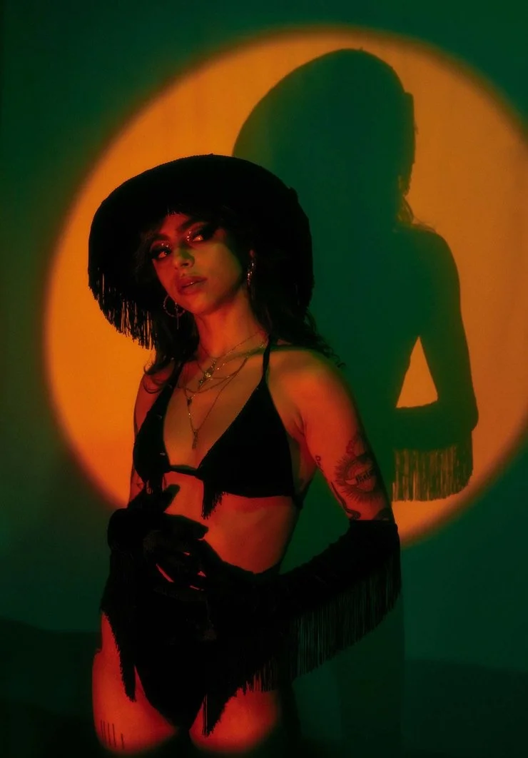 A woman in a black bra with fringe details, wearing long black gloves, stands in front of a colorful circle projection on a wall, with her shadow cast behind her. She has dark hair, layered necklaces, and tattoos on her arms.