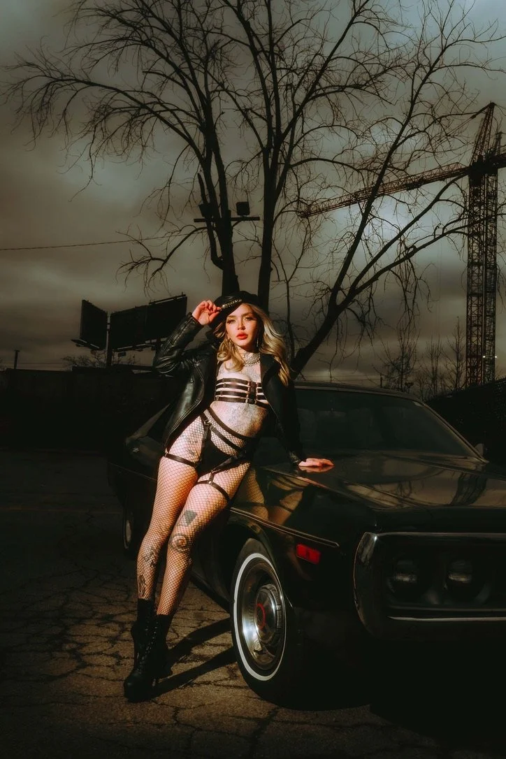 A woman in fishnet stockings, leather jacket, and harness lingerie leaning on a black vintage car, with bare trees and a cloudy sky in the background during dusk.