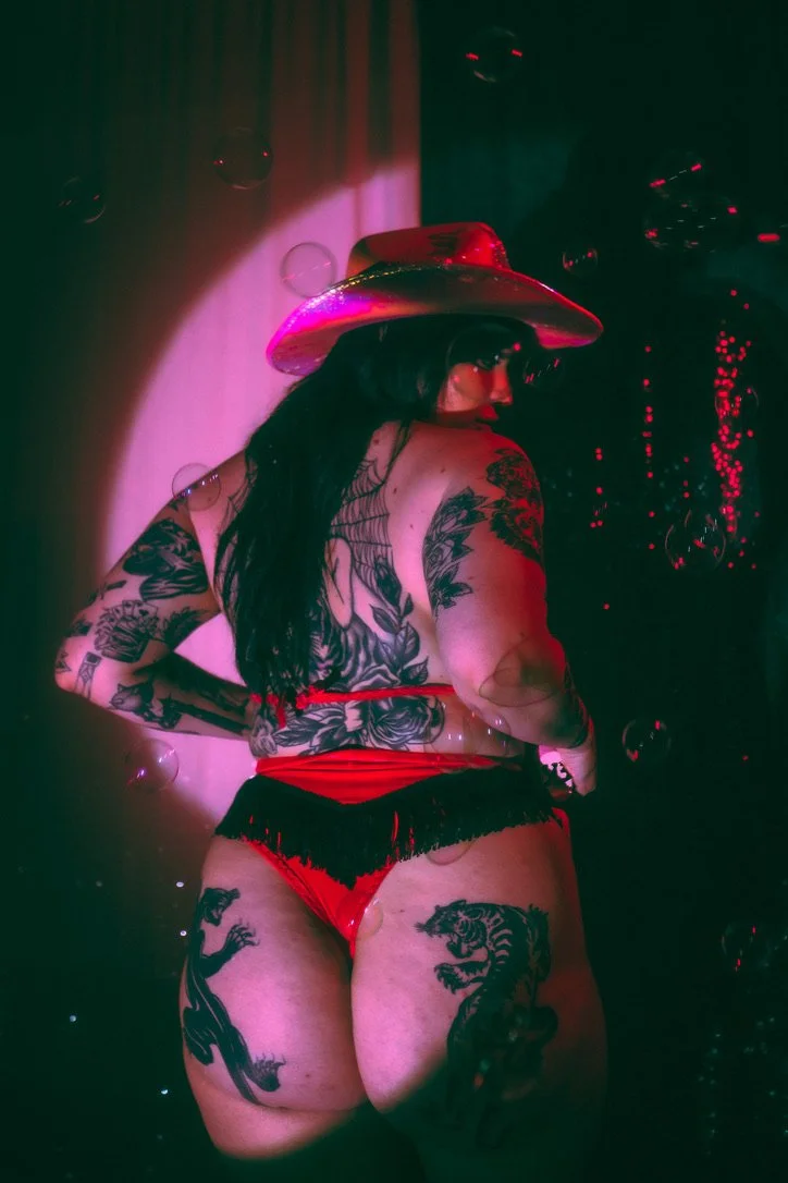 A woman with long dark hair and tattoos on her back and legs, wearing a red hat and red fringe shorts, posing against a dark background with pink and red lighting.