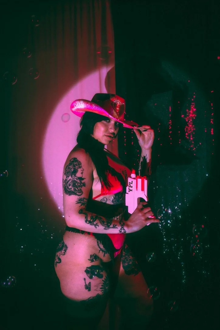 A woman with black hair and tattoos on her arm and legs posing with a toy gun and a pink cowboy hat, illuminated by colorful lights and bubbles.