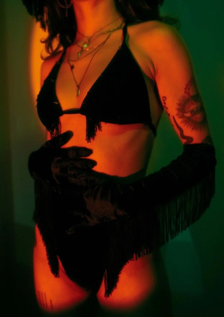 A woman wearing black lingerie and long black gloves, with tattoos on her arm, in a dimly lit room with colored lighting.