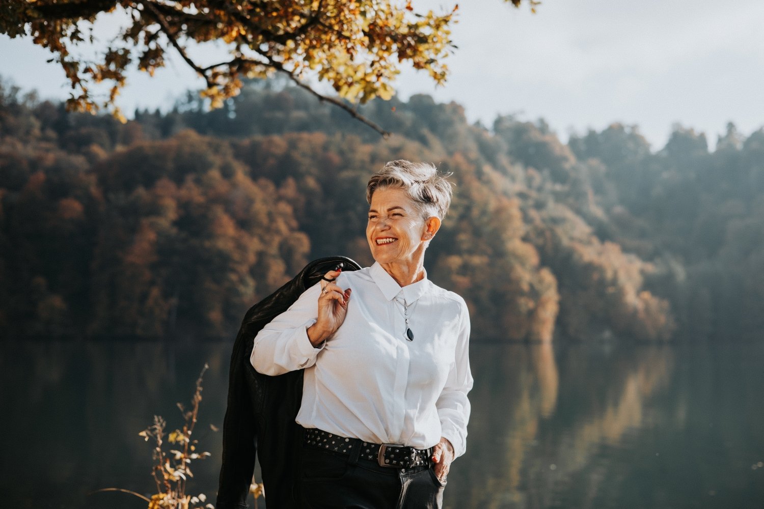 Natürliches Outdoor-Porträt einer Frau am Wasser mit Wald und Bergen im Hintergrund.