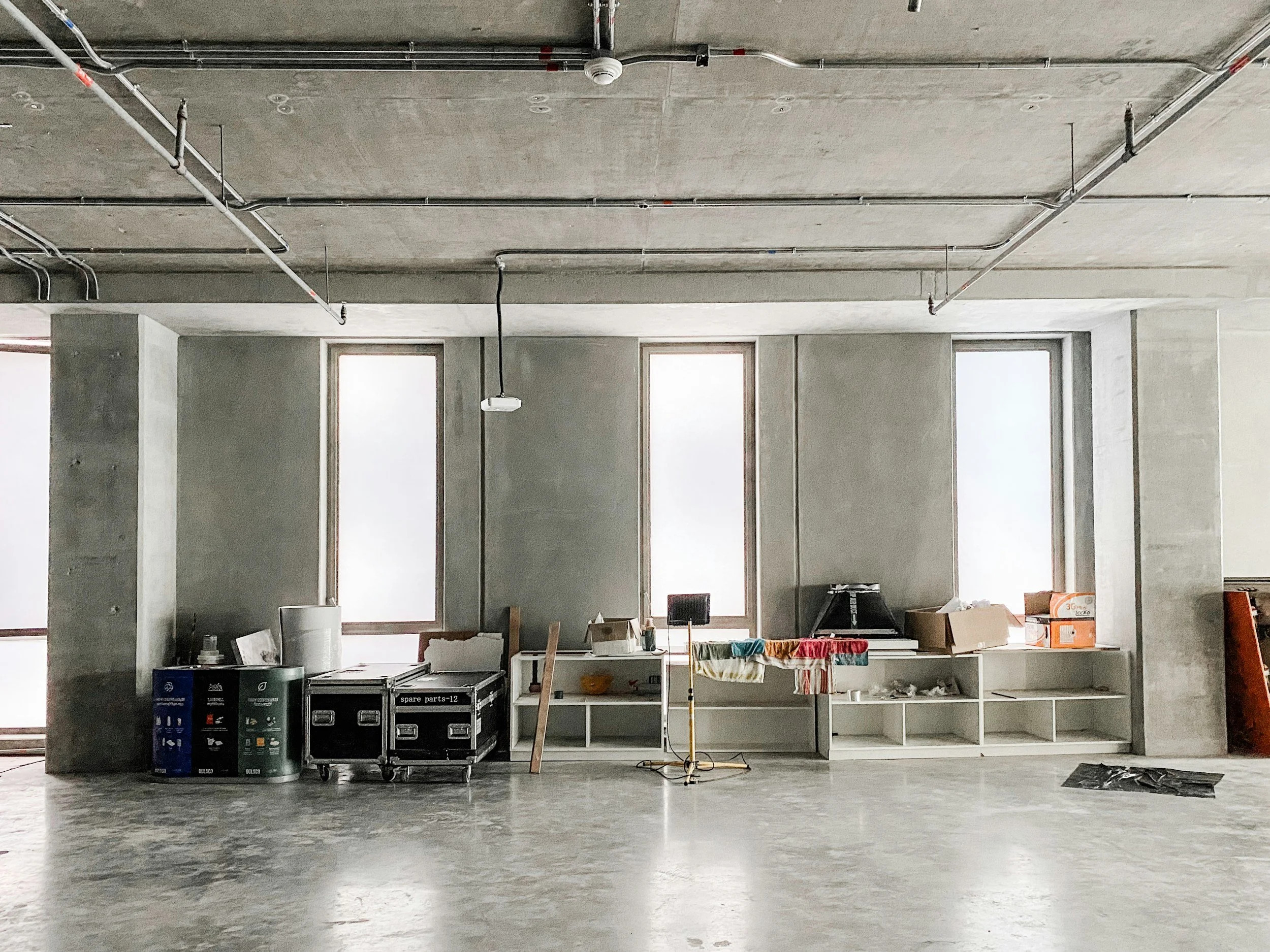 Interior of an unfinished room with large windows, construction materials, storage boxes, and cleaning equipment, indicating ongoing construction or renovation.