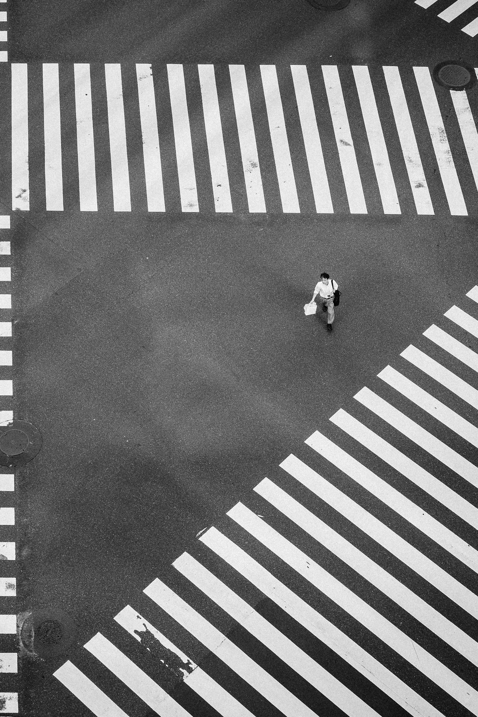 Top-down view of a crosswalk on a street with a person walking across, holding shopping bags and a bag, wearing glasses, a button-up shirt, and pants.