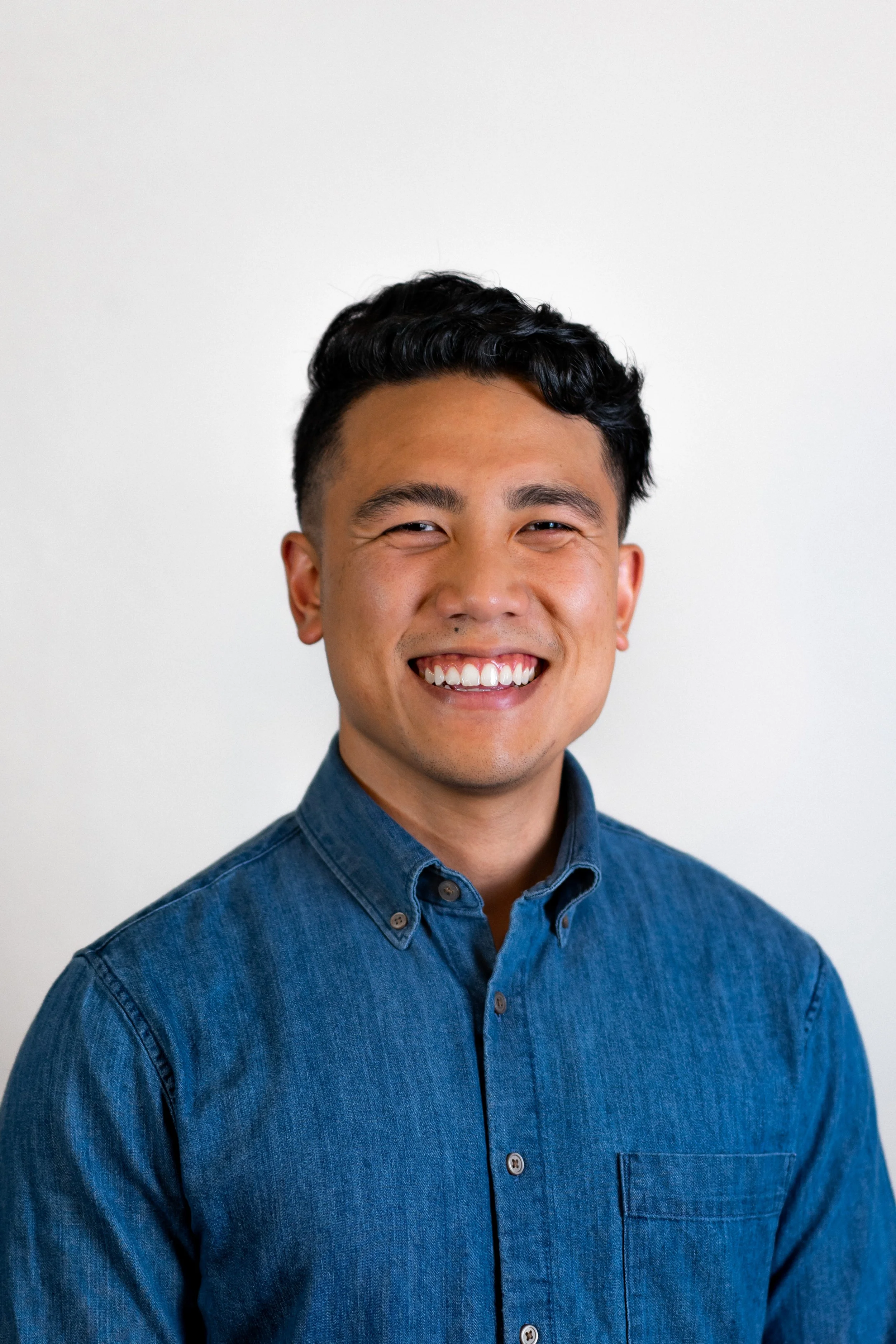Young man with dark hair smiling, wearing a denim button-up shirt, against a plain white background.