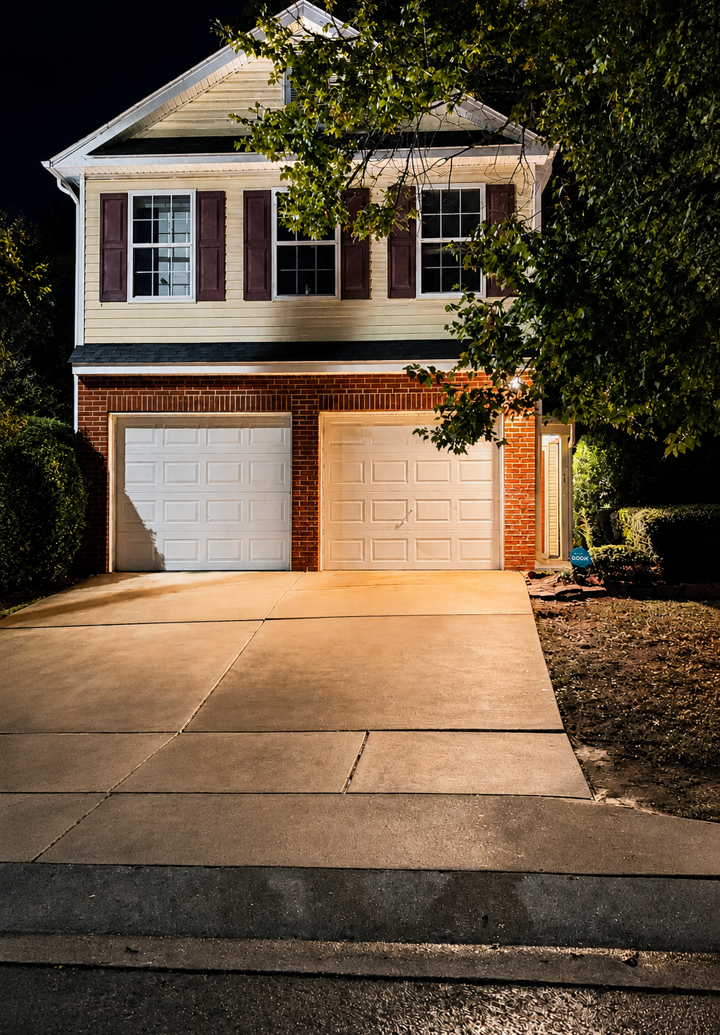 A two-story house at night with a brick and siding exterior. The house has two white garage doors and three windows on the upper floor with purple shutters. There is a driveway and some greenery, including a large tree partially obscuring the house.