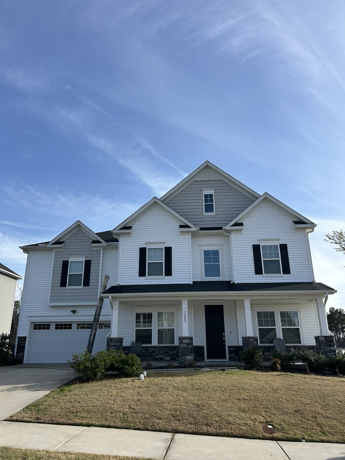 Front view of a modern three-story white house with black shutters, stone accents, and a two-car garage, under a blue sky with wispy clouds.