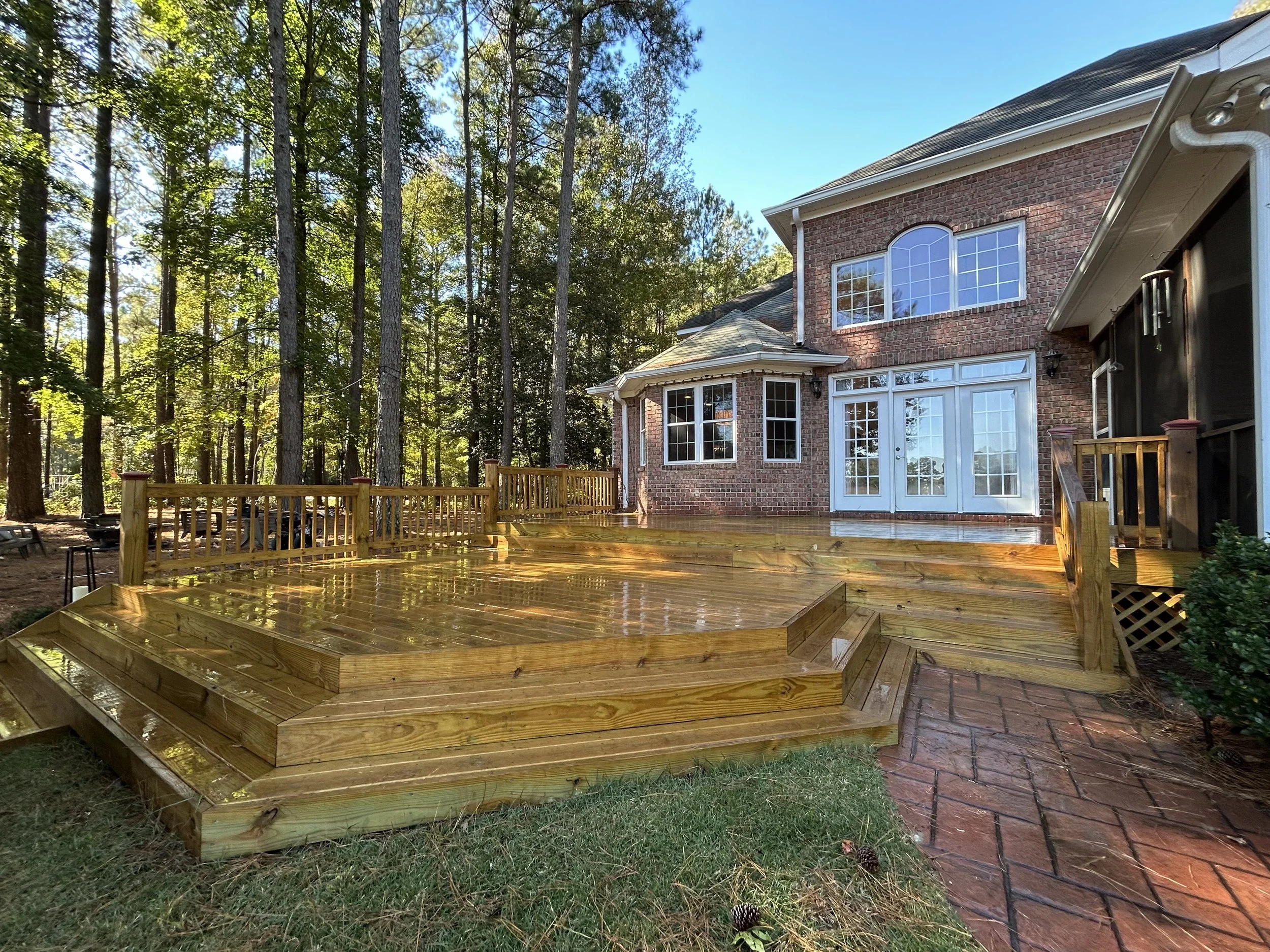 A backyard with a wooden deck attached to a brick two-story house surrounded by tall trees and a bright blue sky.