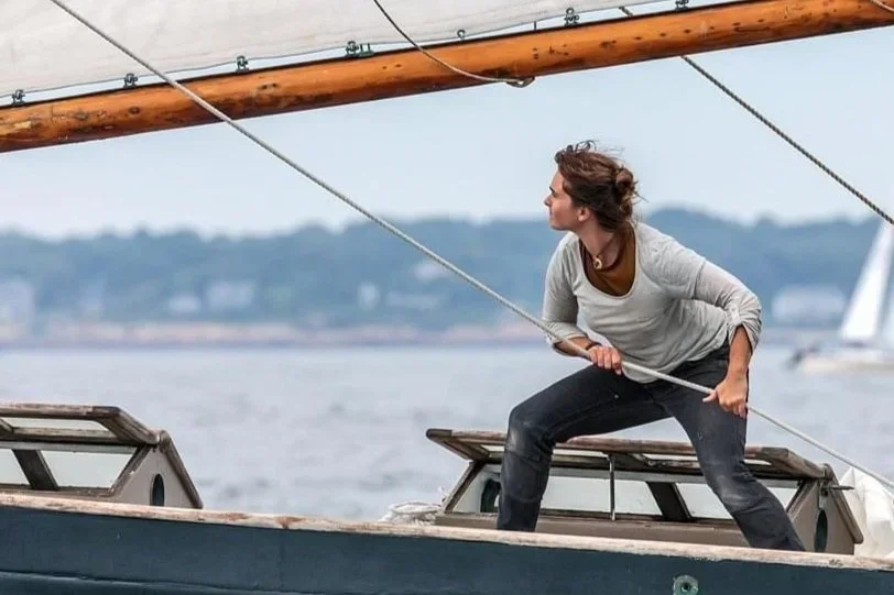 A woman is on a sailboat, leaning forward and pulling a rope, with water and land in the background.