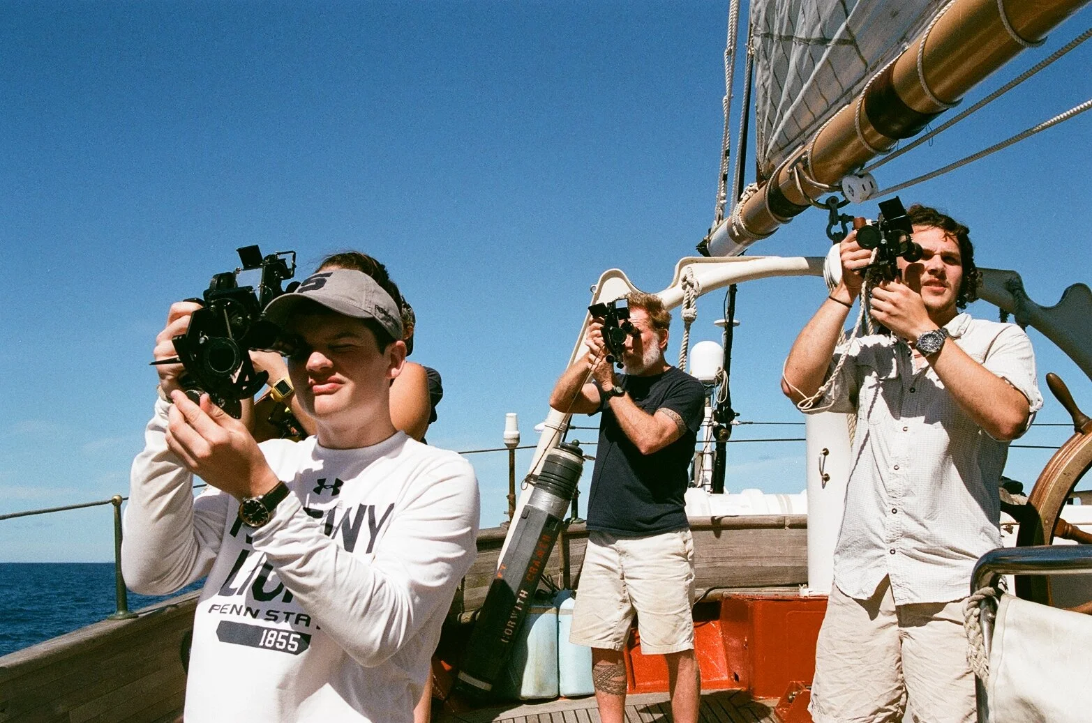 Three men on a boat with one holding a camera, the other pointing binoculars, and the third looking through a camera, with clear blue sky and ocean in the background.