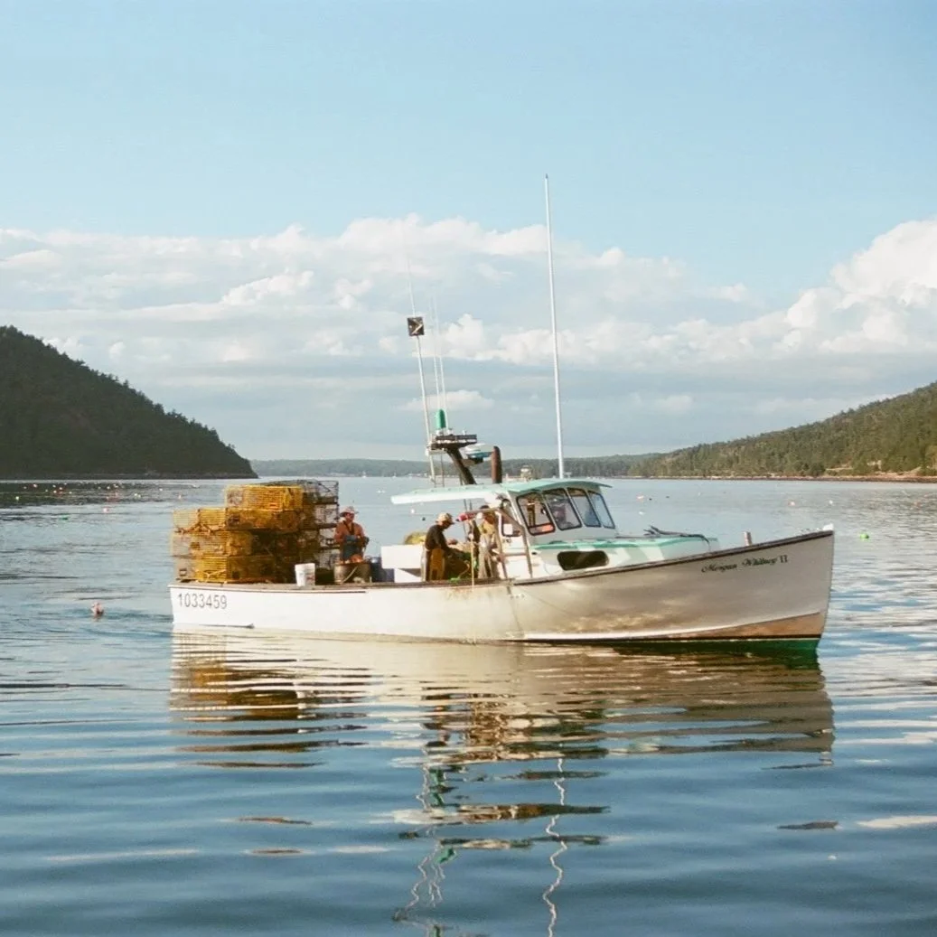 A boat on a calm lake with a clear sky, surrounded by hills, carrying stacks of lobster traps and three people standing on deck.
