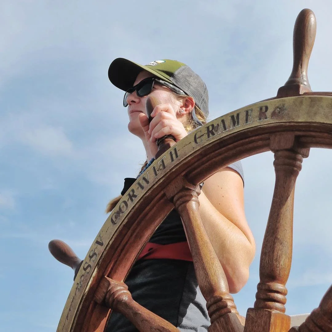 A woman wearing sunglasses and a cap steering a ship's wheel with the words "Torngat" and "Graham" engraved on it, against a background of a cloudy sky.