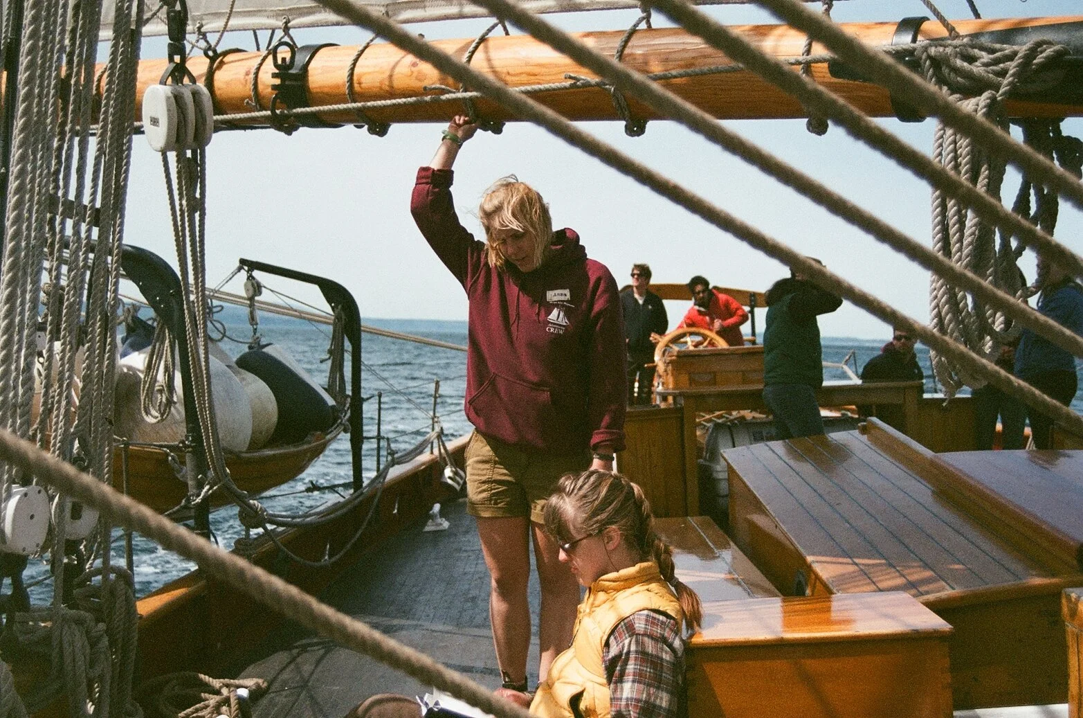 Crew members working on a wooden sailboat in the open sea during daytime, with ropes and equipment visible, and a clear sky in the background.