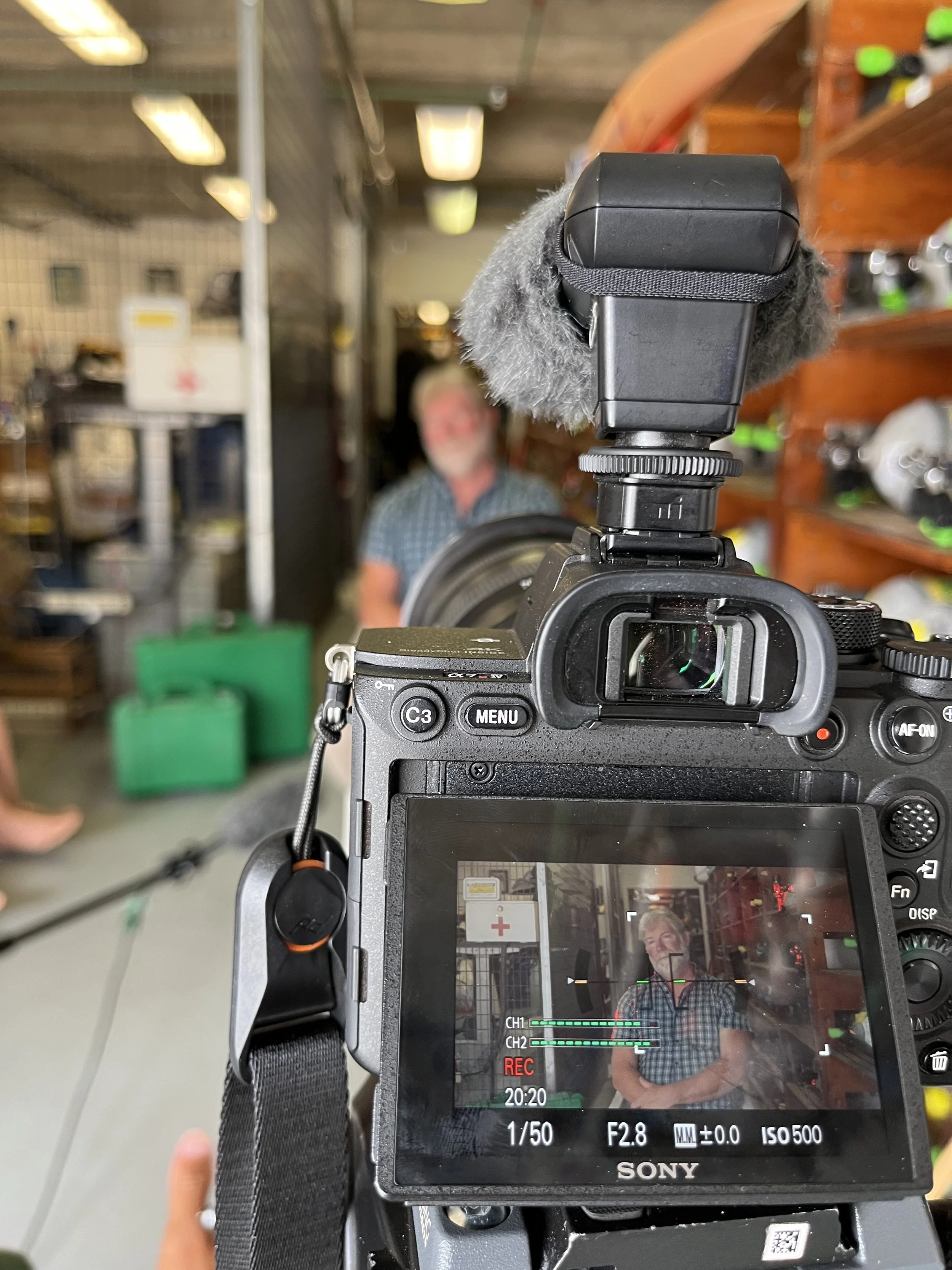 The image shows the view through a camera's LCD screen. In the background, a man with gray hair and a checkered shirt stands inside a store, possibly a hardware or sporting goods store. The camera's mounted microphone is covered with a furry windscreen. The store has shelves with various products, and fluorescent lighting is visible on the ceiling.