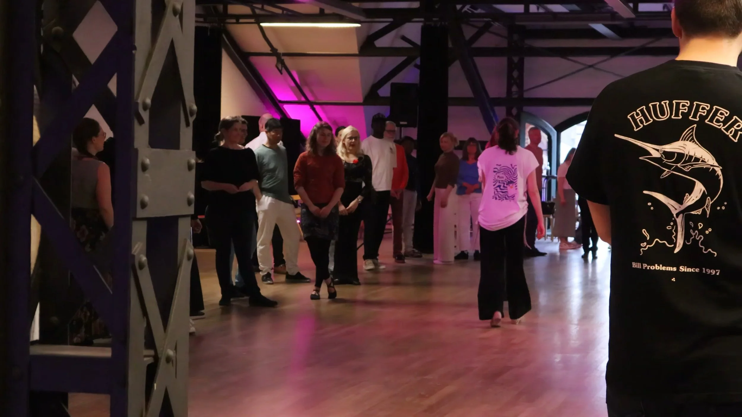 Group of people standing and walking in a dimly lit indoor space with pink and purple lighting, possibly at an event or gathering.