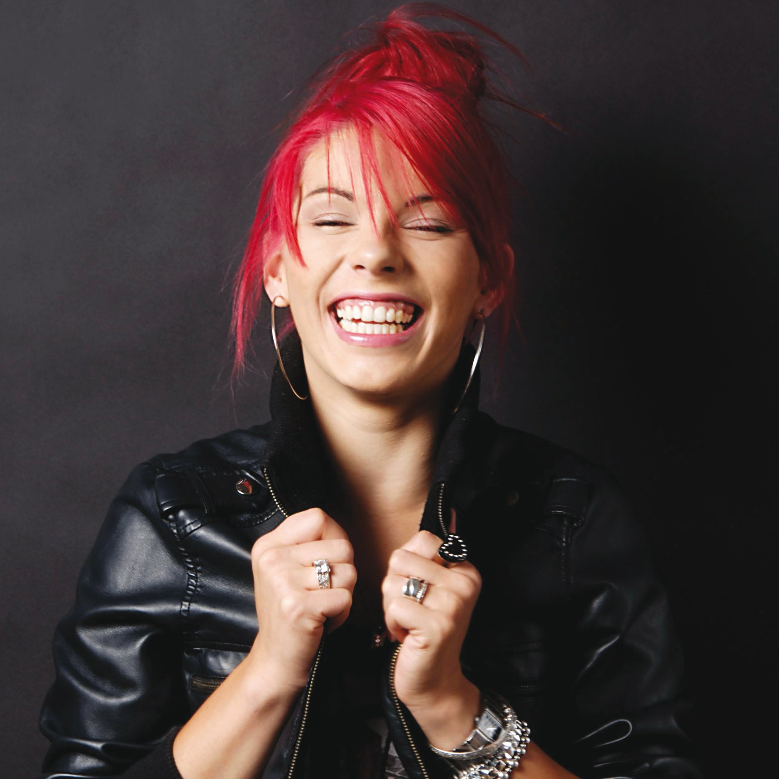 A woman with bright red hair styled in a spiky manner, smiling with closed eyes and showing teeth, wearing hoop earrings, multiple rings, a leather jacket, and a black shirt, against a dark background.