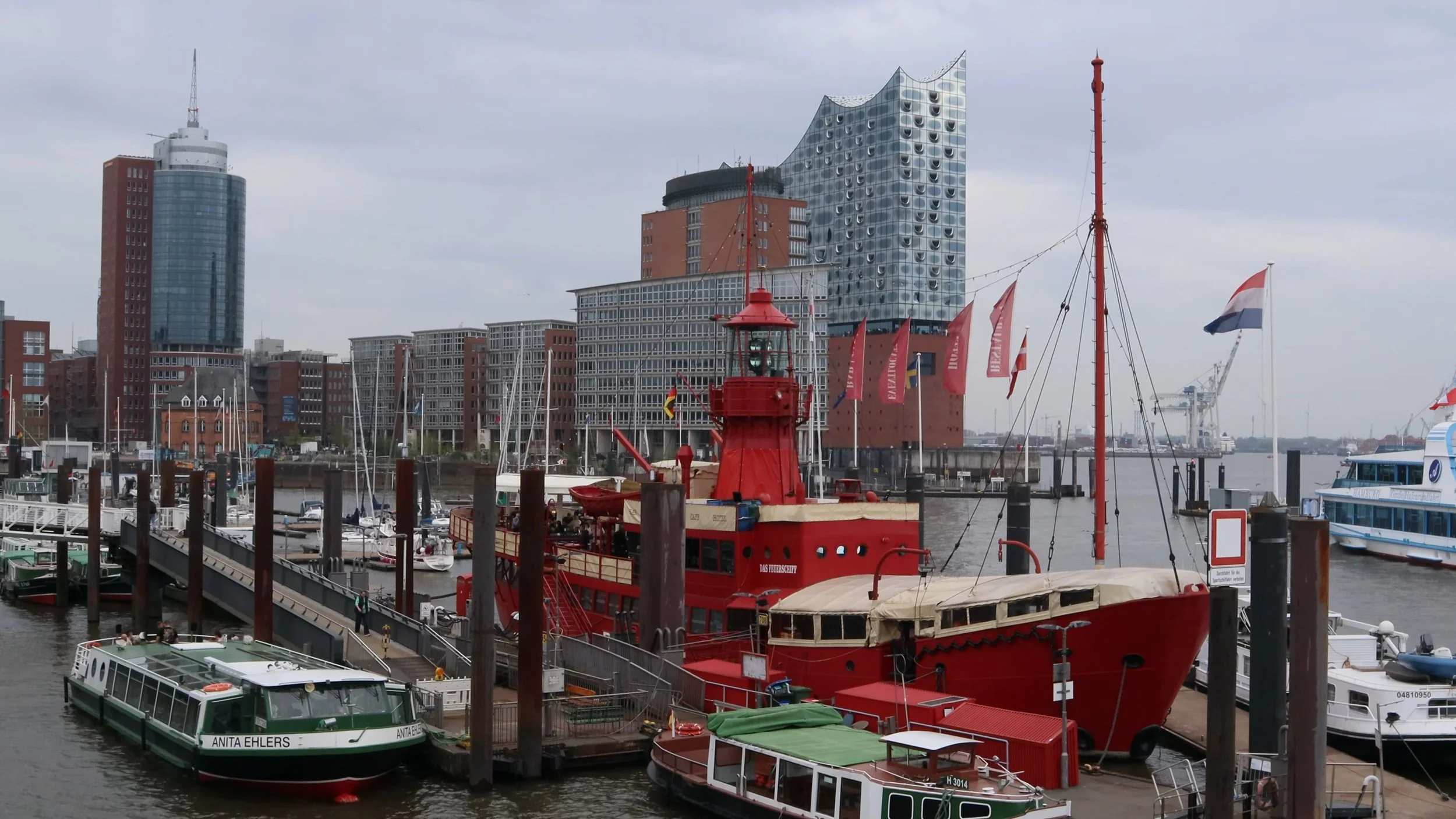 View of a harbor with boats docked, including a prominent red rescue boat, and a city skyline with modern buildings in the background.