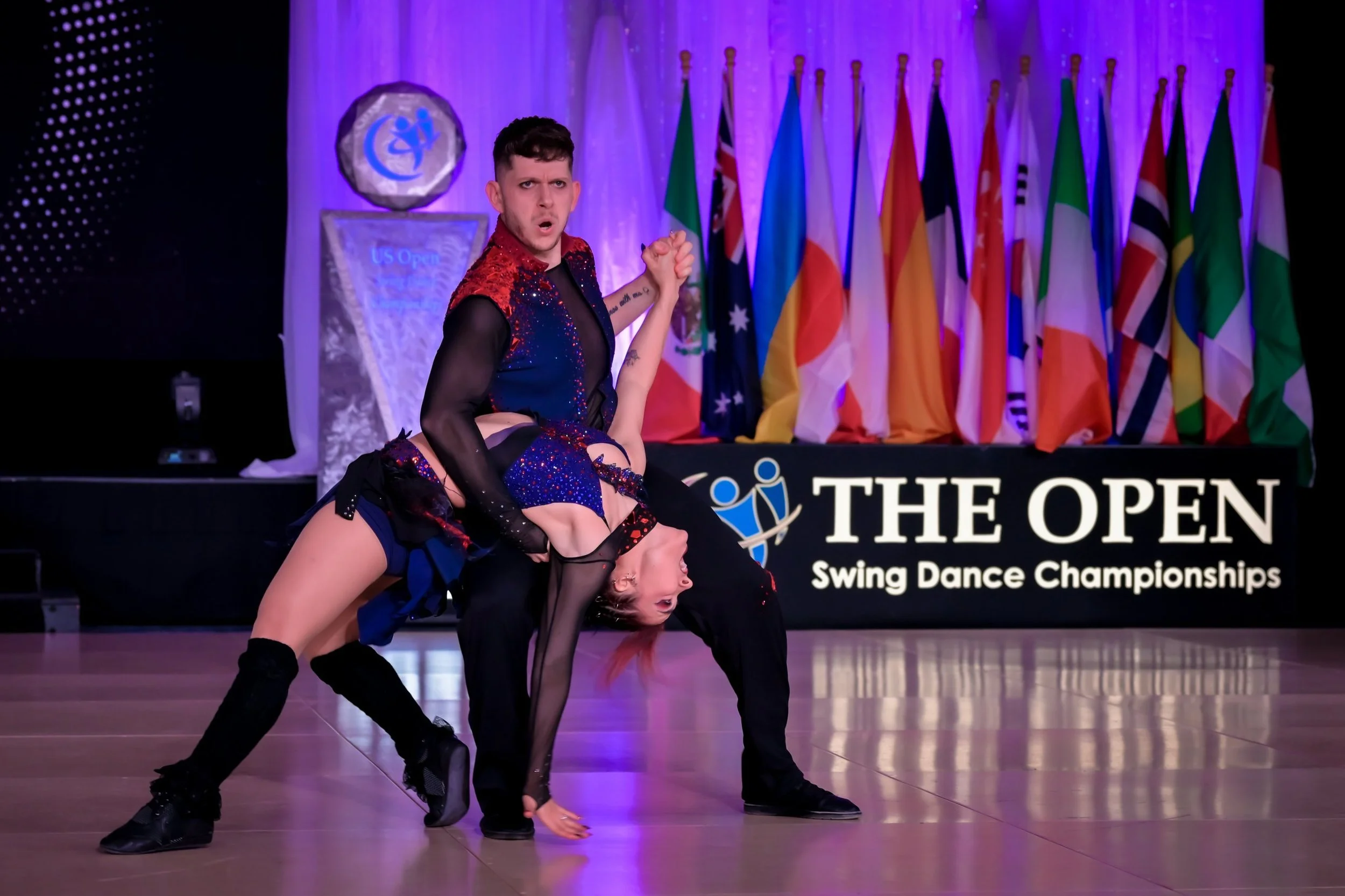 Two dancers performing a swing dance routine on a stage at the US Open Swing Dance Championships. The male dancer is wearing a black and red outfit, and the female dancer is in a black dress with blue accents. There are multiple international flags in the background.