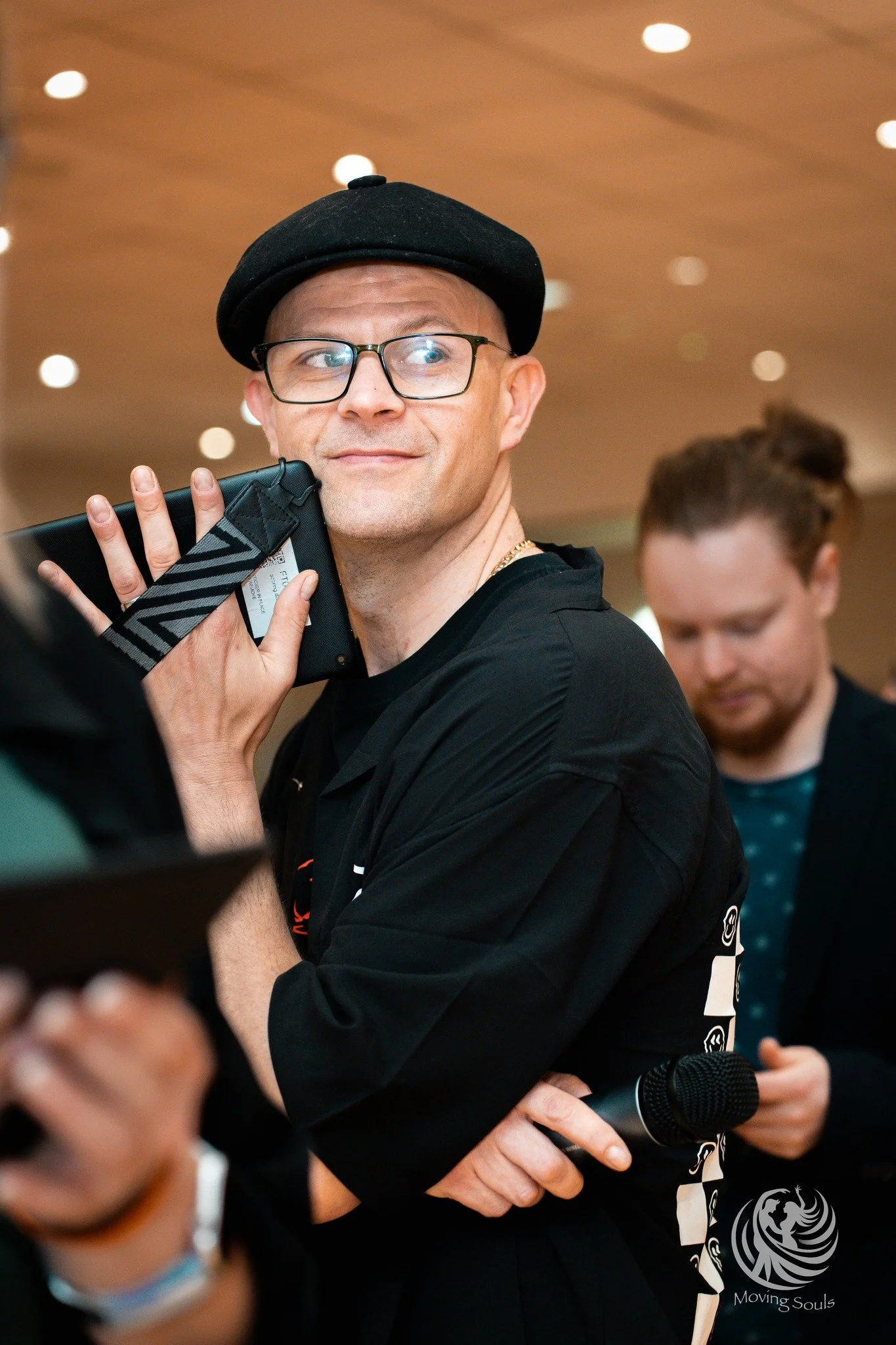A man wearing glasses and a black cap holds a smartphone with a patterned strap close to his ear, smiling slightly. There is a woman in the background and a microphone in front of him, indicating a public or media event.