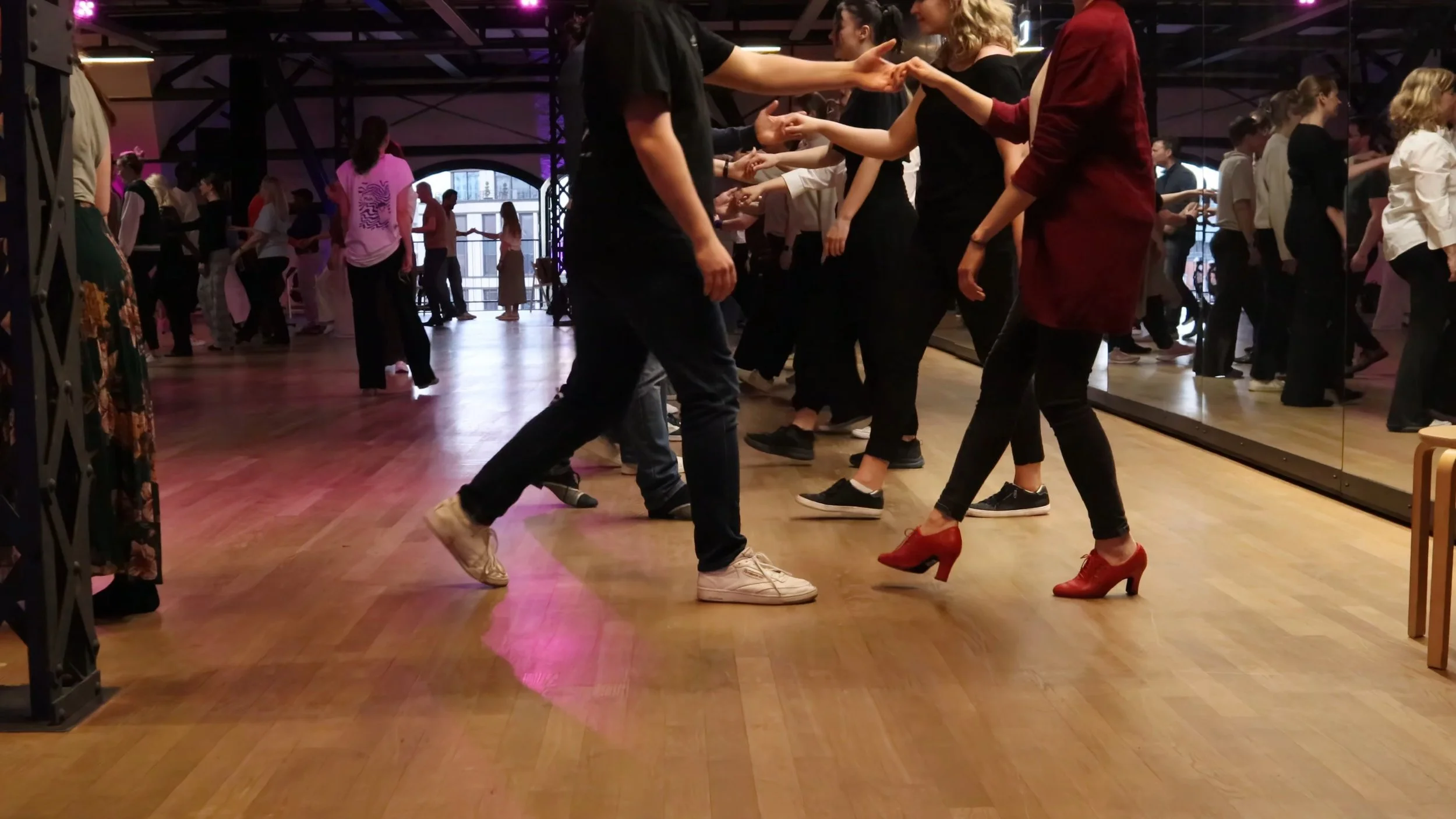Couple dancing in a dance studio with a mirrored wall, surrounded by other dancers, some wearing dance shoes, with colorful lighting.