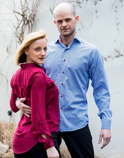 A man and woman standing close together outdoors on a cloudy day, with the woman wearing a red satin blouse and the man in a blue checkered shirt.