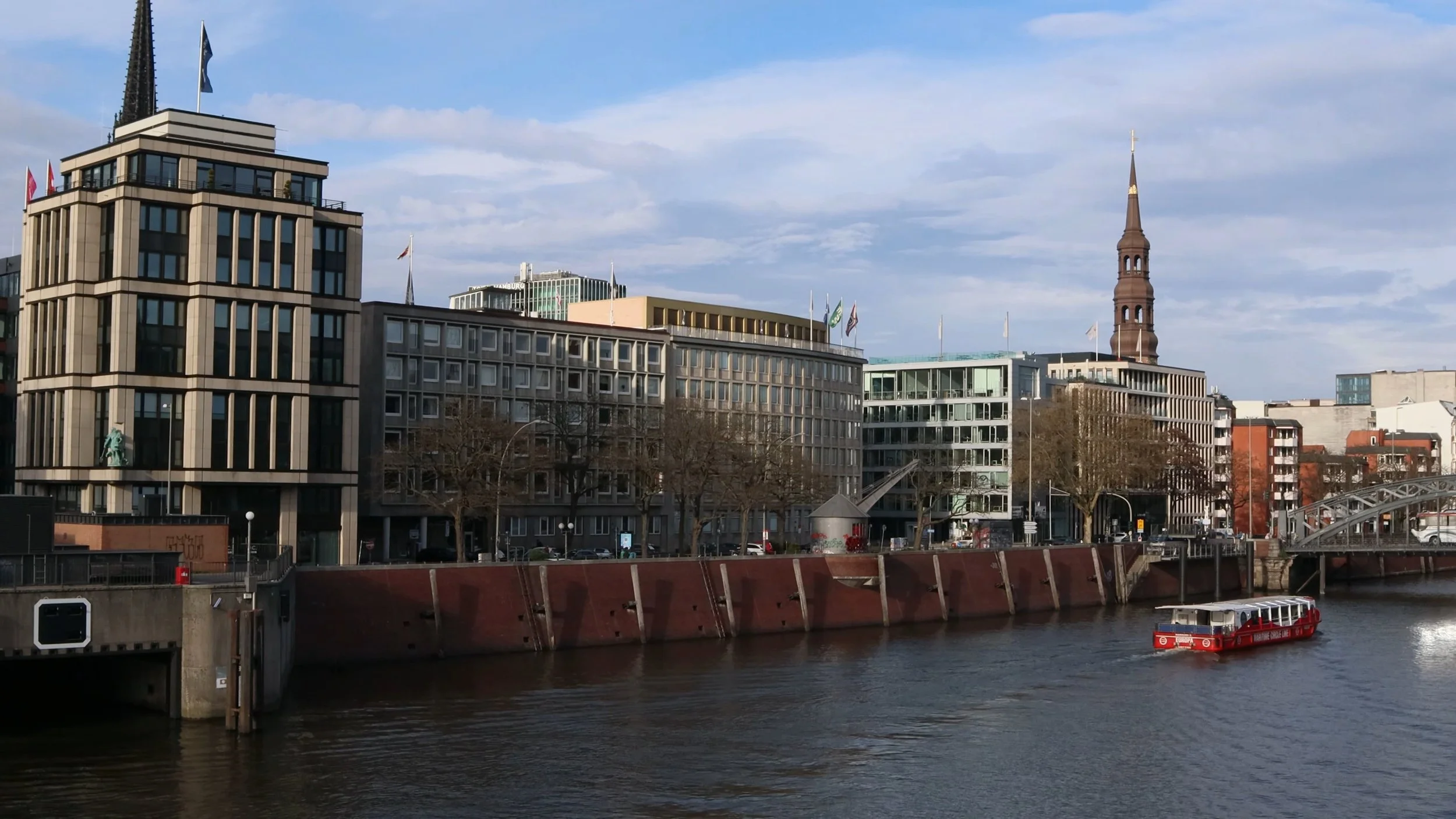 Cityscape with modern buildings along a river, including a church steeple, a boat on the water, and a bridge.