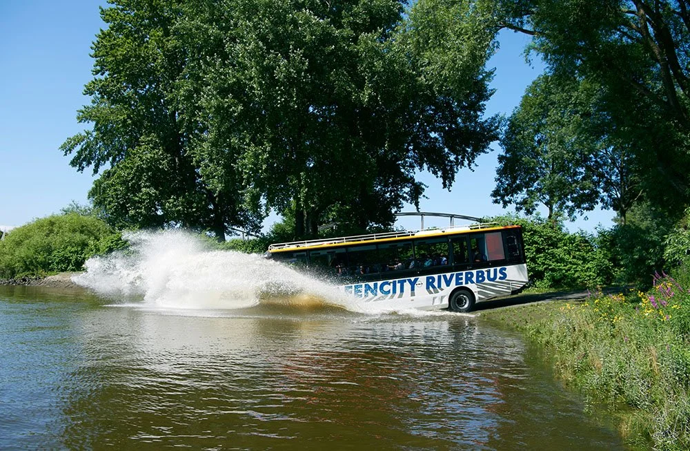 A city river bus labeled 'FENCITY RIVERBUS' is traveling on the water, creating a splash as it speeds past the shoreline with trees and grass.