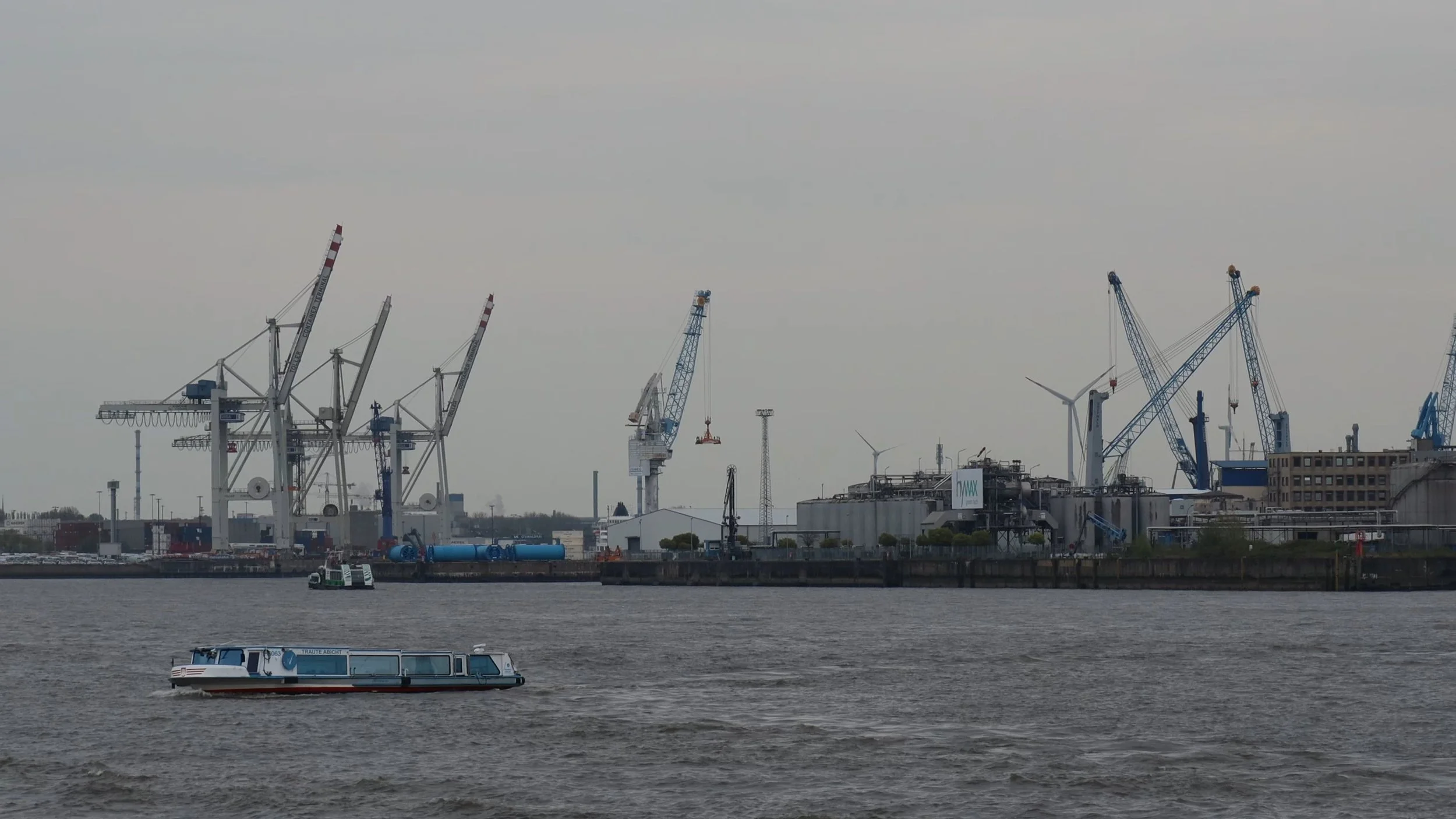 A harbor scene with multiple cranes, wind turbines, and industrial buildings, viewed across a body of water with a boat in the foreground.