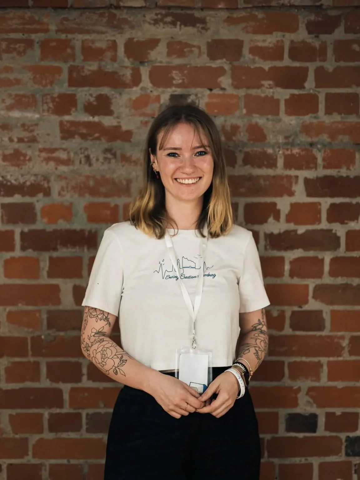 A woman with shoulder-length blonde hair, blue eyes, and tattoos on her arms standing against a plain white wall. She is smiling, wearing a black sleeveless top and jewelry, with her hands on her hips.