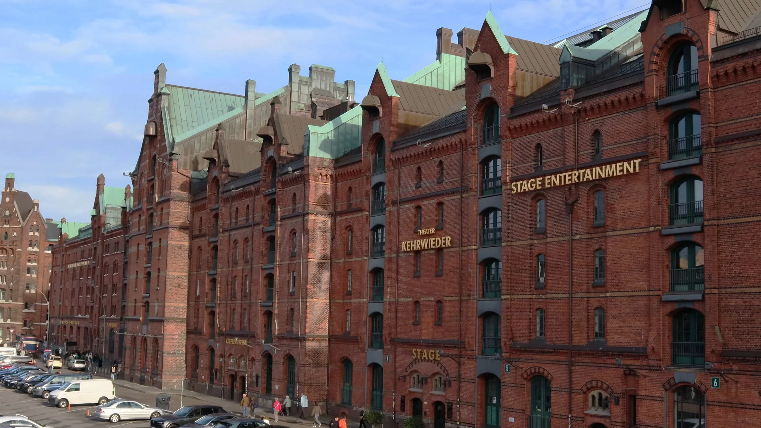 A large red brick building with multiple stories and green rooftops, labeled with signs such as 'Stage Entertainment' and 'Kehrwieder,' in a city setting with parked cars and pedestrians.