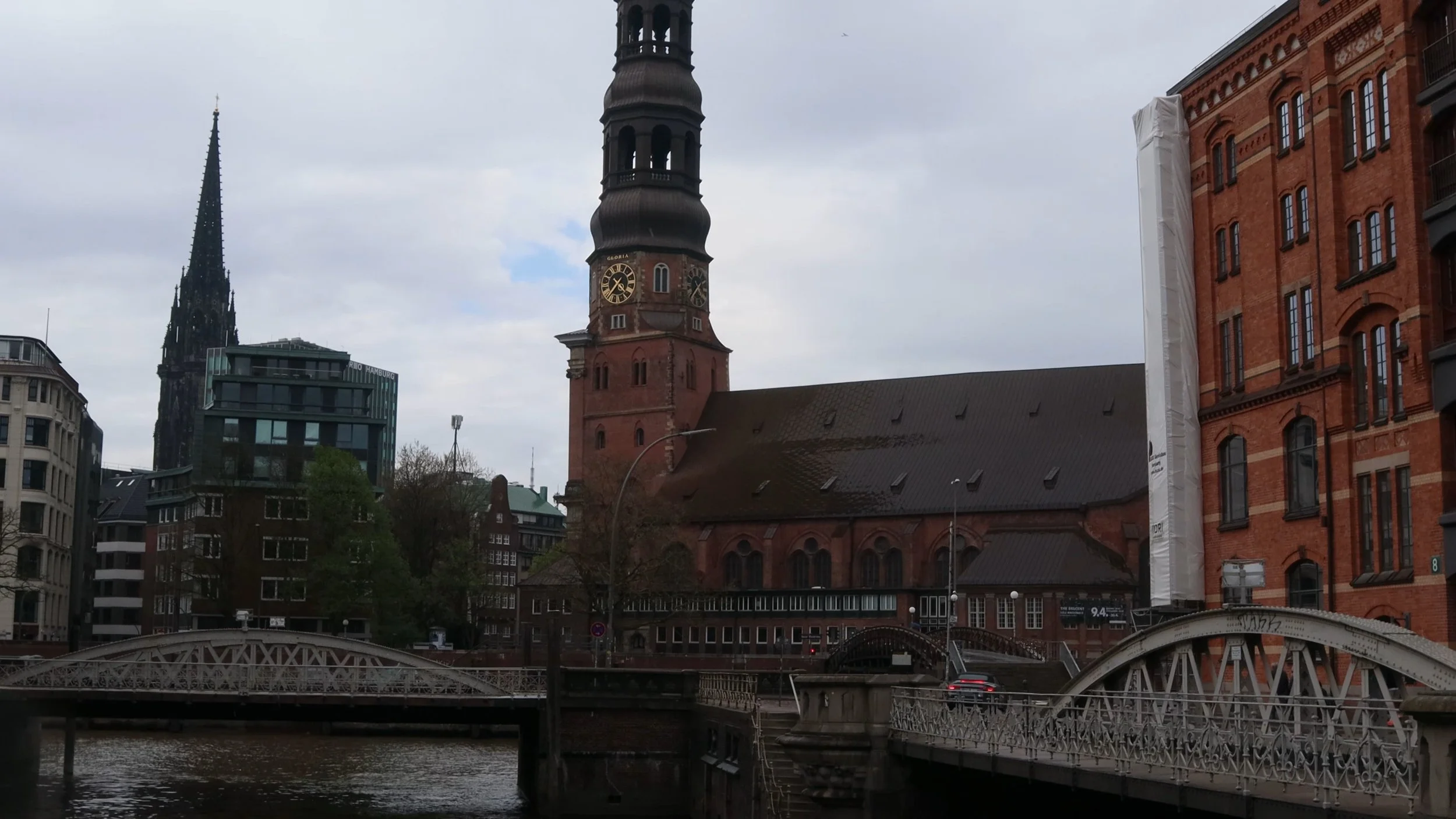 Historical church with a tall clock tower on a cloudy day, surrounded by modern buildings and a bridge over a river.