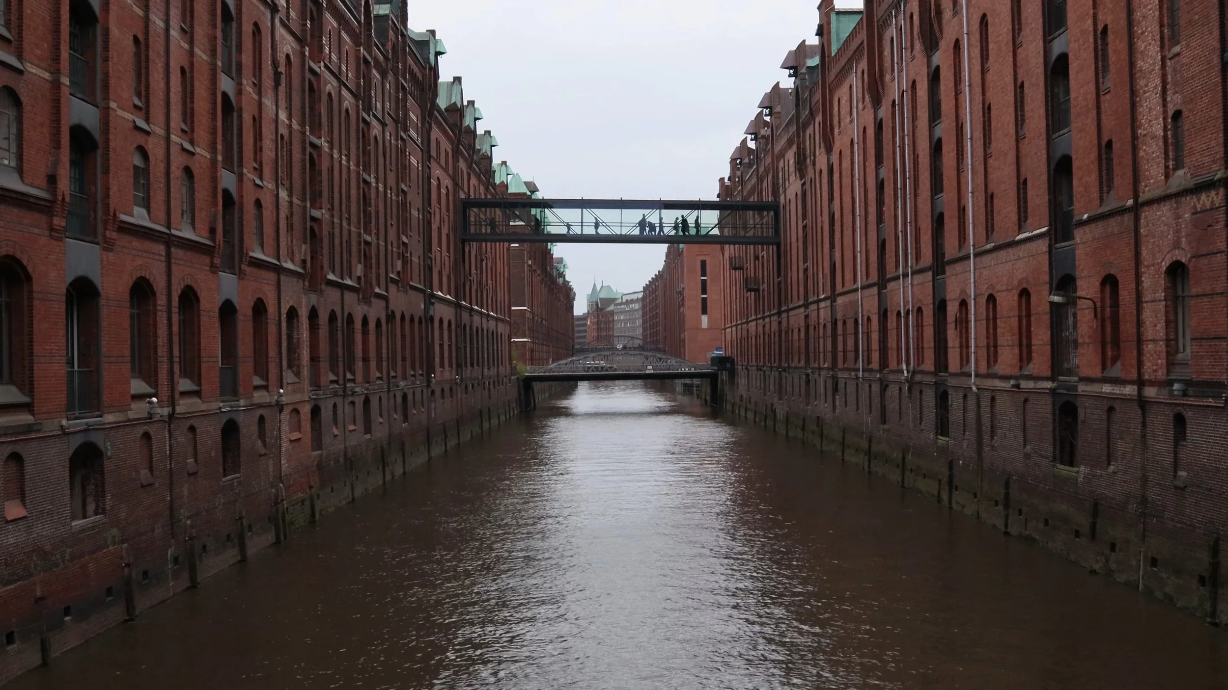 Red brick warehouses lining a canal with footbridges and people walking on a skywalk above the water
