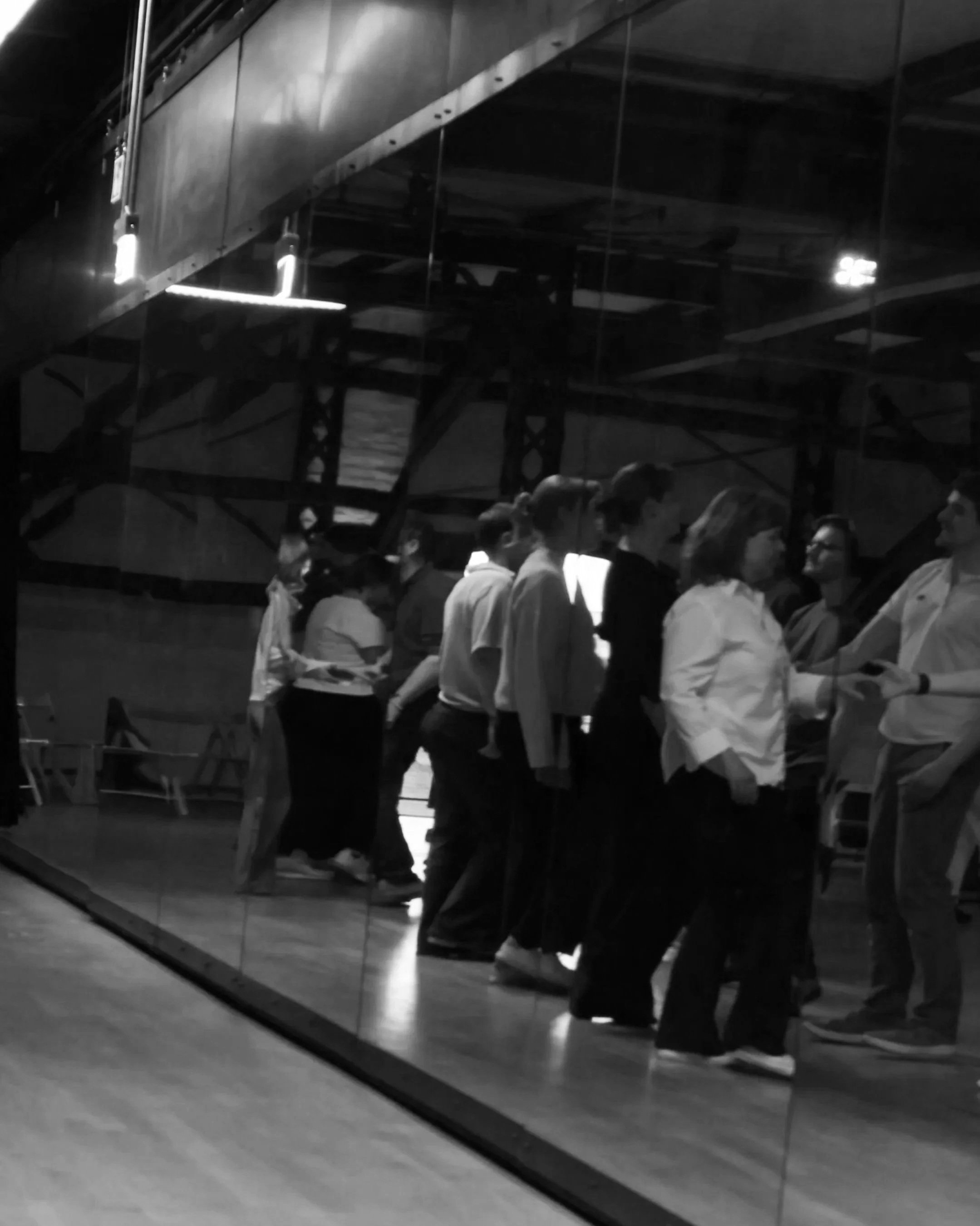 A group of people walking inside a building, reflected in glass walls, with some wearing glasses and long hair, possibly in an urban or modern environment.