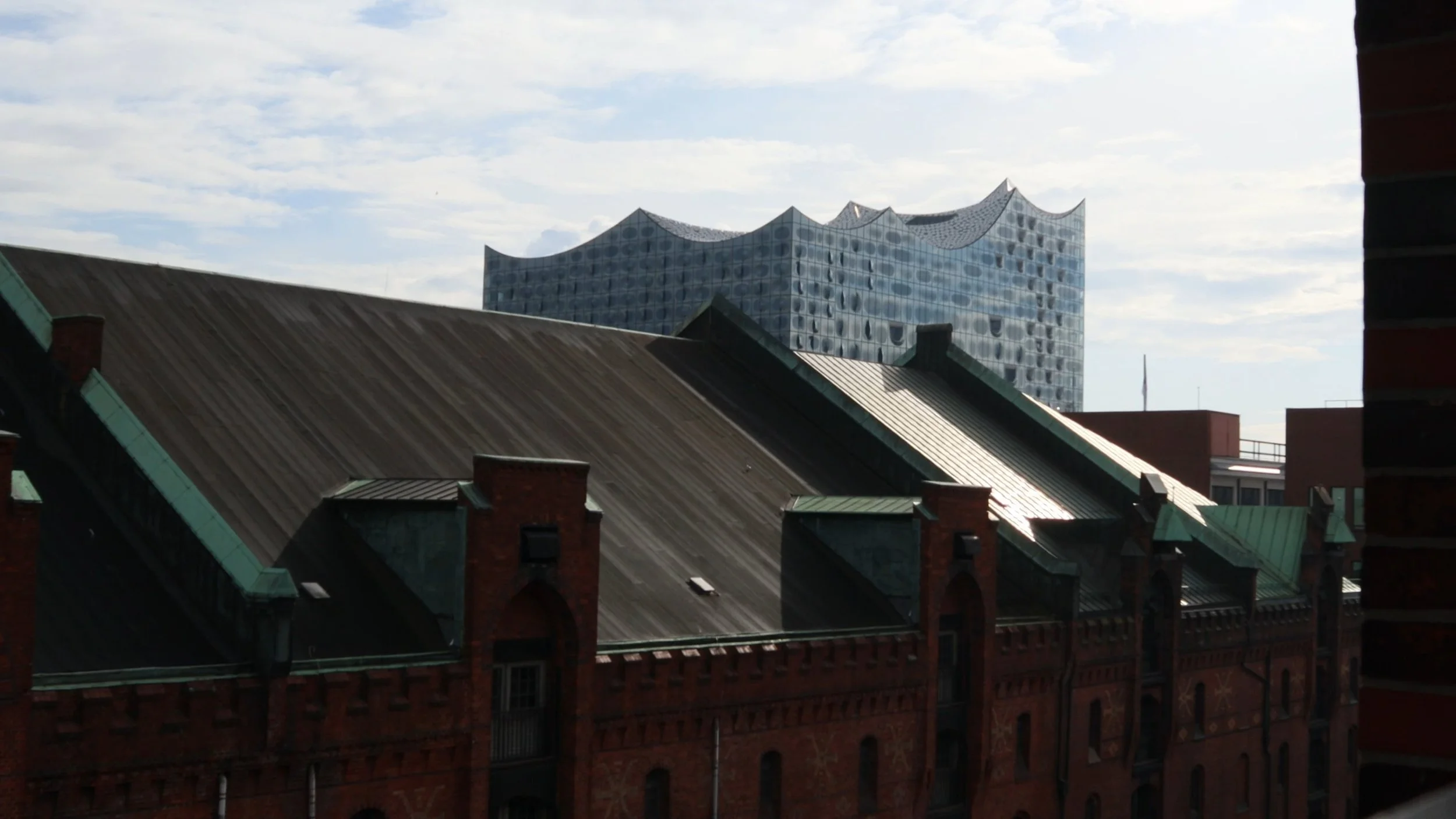 ELBPHILHARMONIE  - A modern building with a wavy, glass facade in the background, viewed behind older, traditional brick buildings with pitched roofs in the foreground.