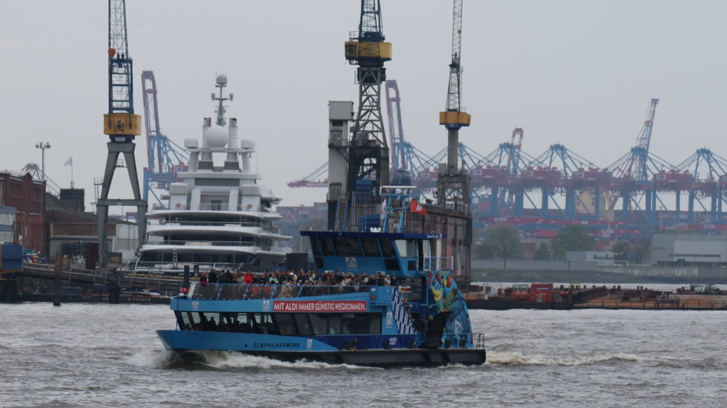 A blue sightseeing boat named 'Elbphilharmonie' on the river with passengers onboard, sailing past a dock with industrial cranes and large ships in the background.