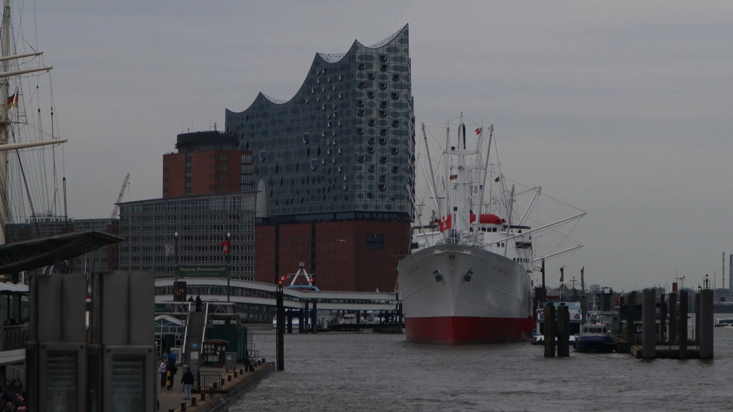A view of a marina with parked boats, including a large ship in the foreground, and modern buildings in the background under an overcast sky.