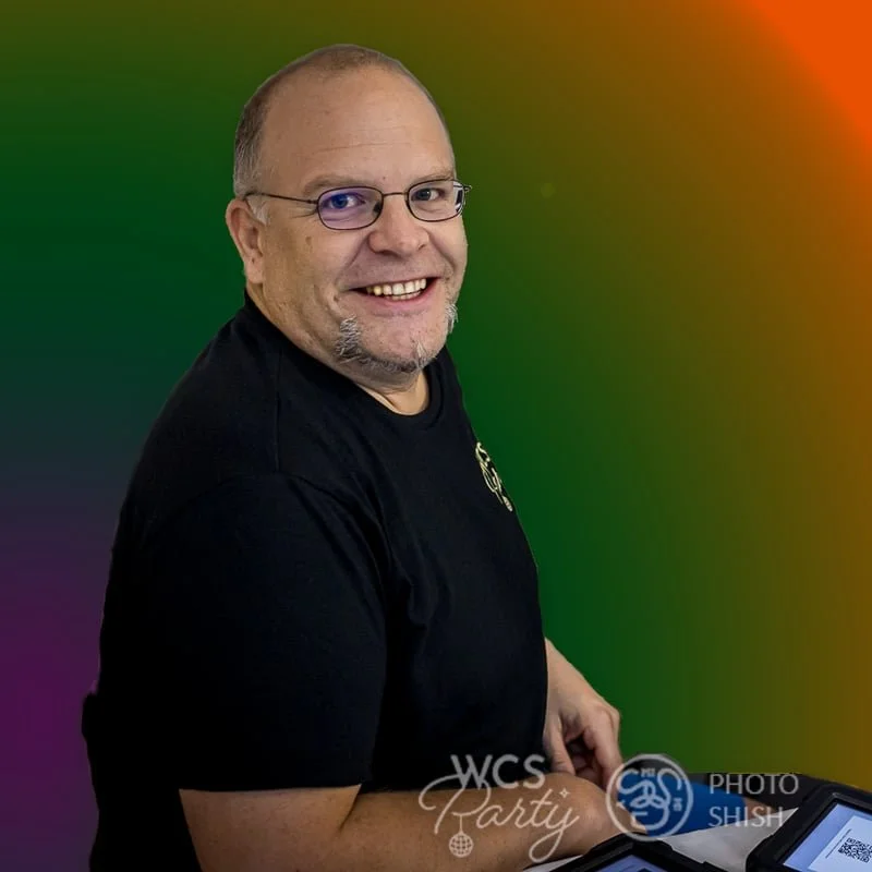 A smiling man with glasses and a goatee sits at a table, wearing a black shirt with a logo, with a colorful gradient background.
