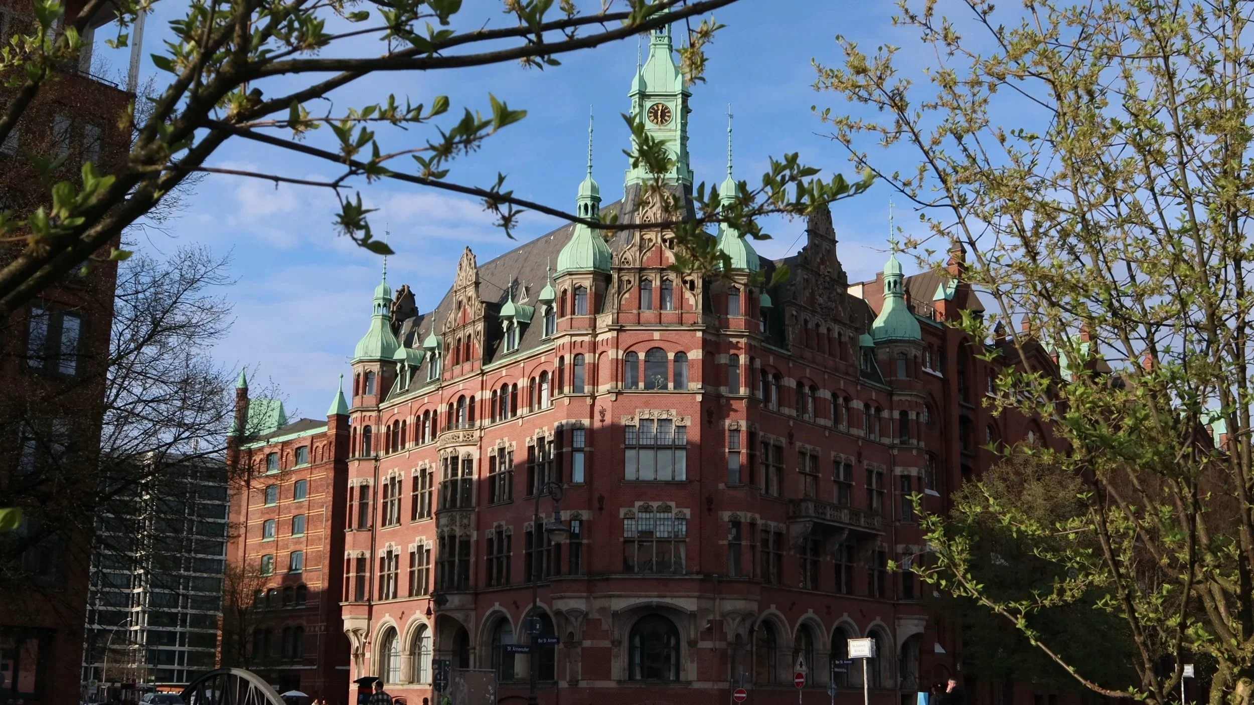 A historic red-brick building with multiple green copper spires and a clock tower, partially framed by trees with budding leaves, under a blue sky.