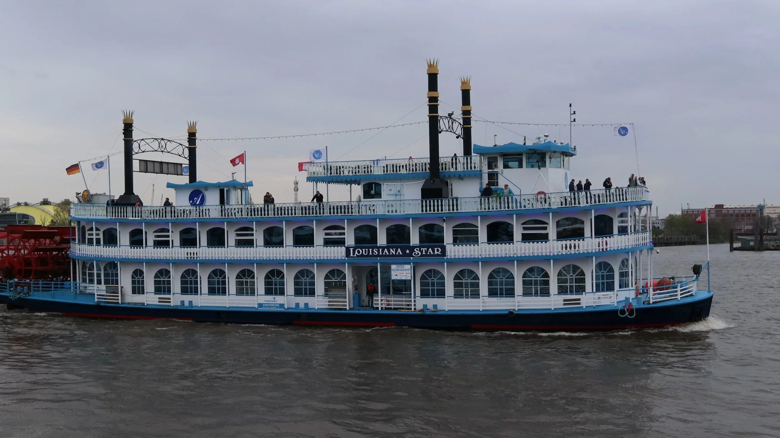 A large, multi-tiered paddlewheel riverboat named Louisiana Star, painted blue and white, sailing on a river with passengers on the upper deck. The boat has two tall black smokestacks with decorative gold tops, and flags, including the German flag an