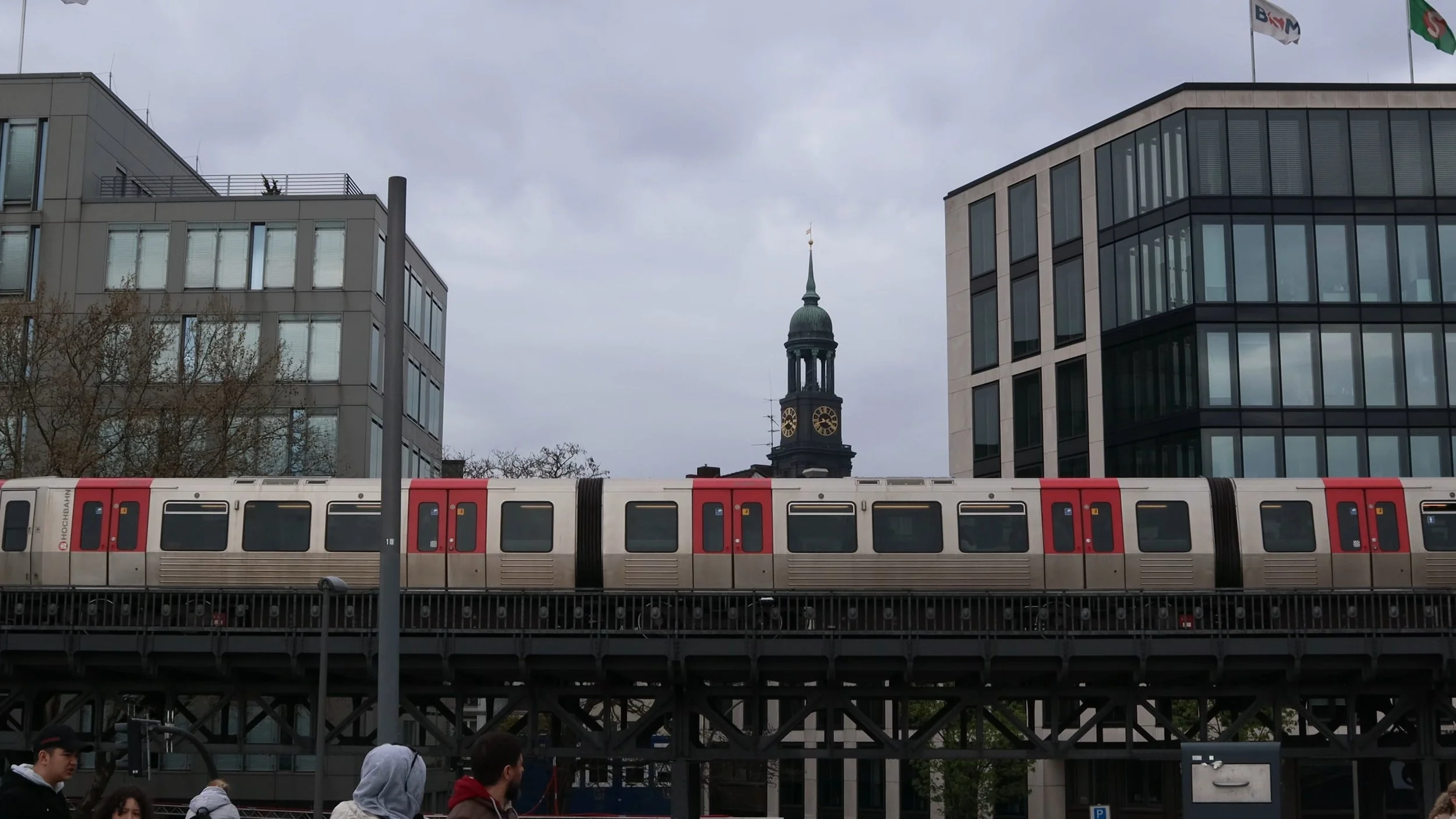 City street scene with modern office buildings, a train on an elevated track, and a historic clock tower in the background under a cloudy sky, with pedestrians walking below.