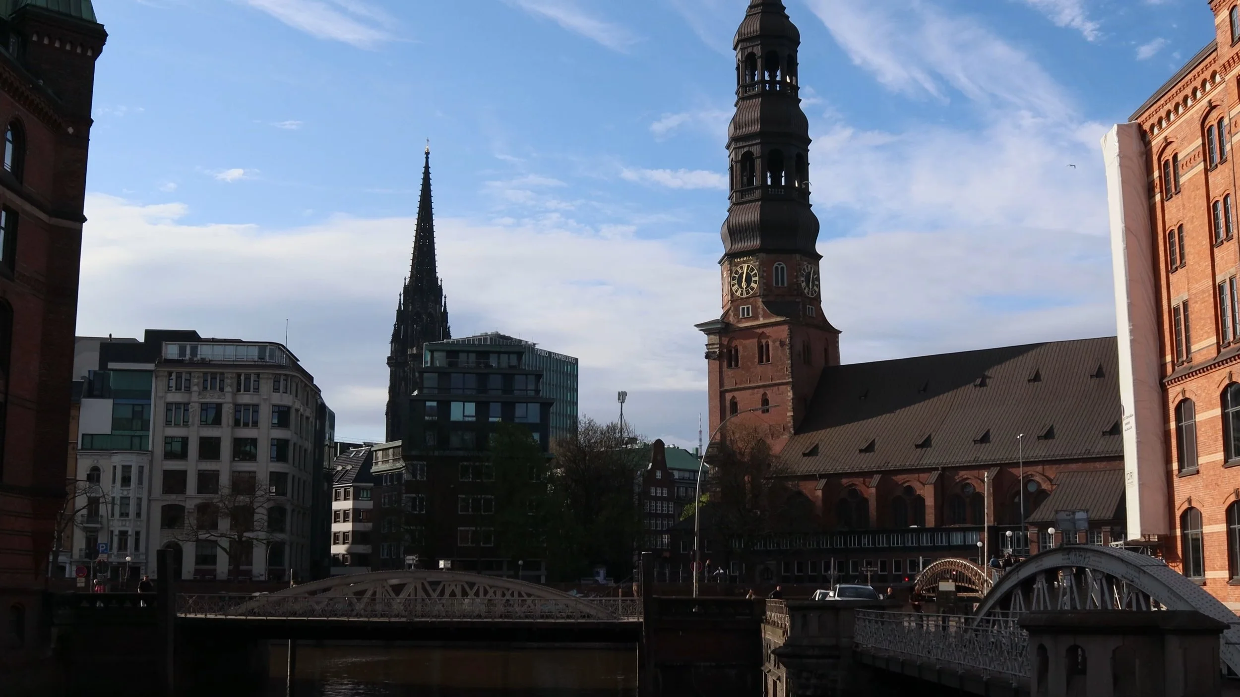 View of historic church and modern buildings along a river in a city, with a bridge in the foreground and a partly cloudy sky.